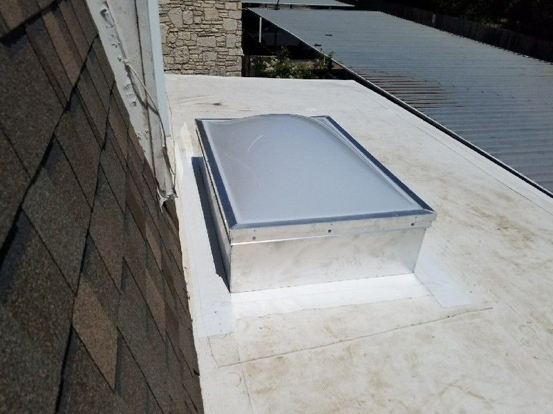 Skylight on a white flat roof, beside brown shingled roof.