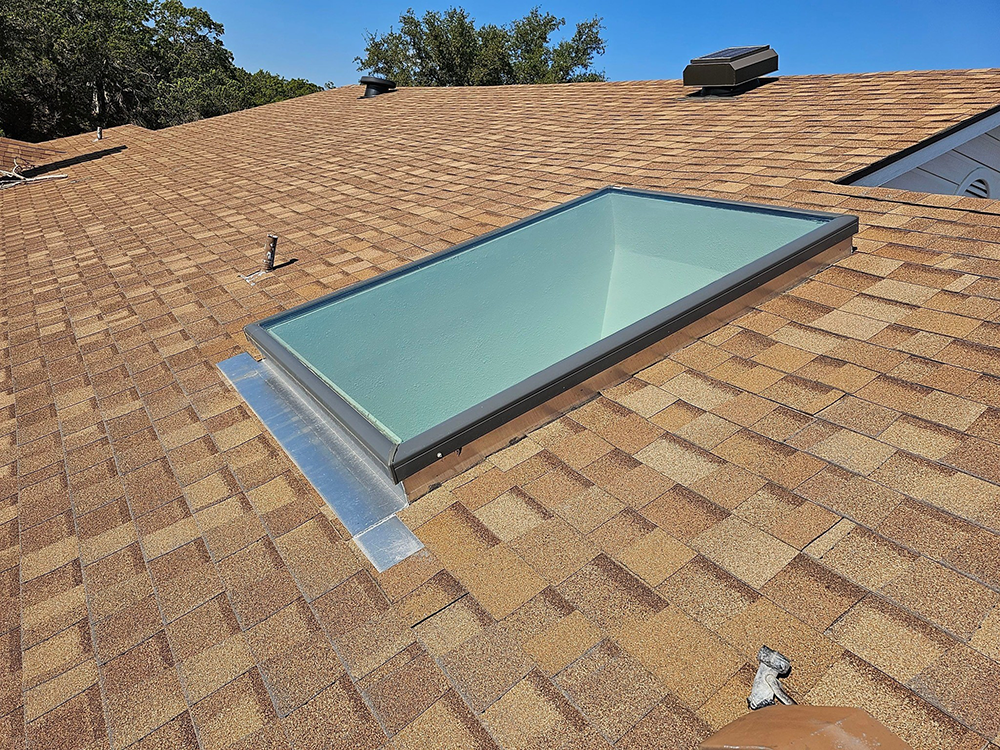 Skylight on a brown shingled roof with a blue interior, on a sunny day.