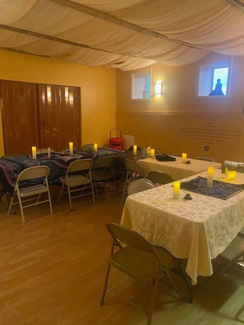 Room set up for an event. Tables with candles and chairs arranged on wood floor, cream-colored walls, cloth ceiling.