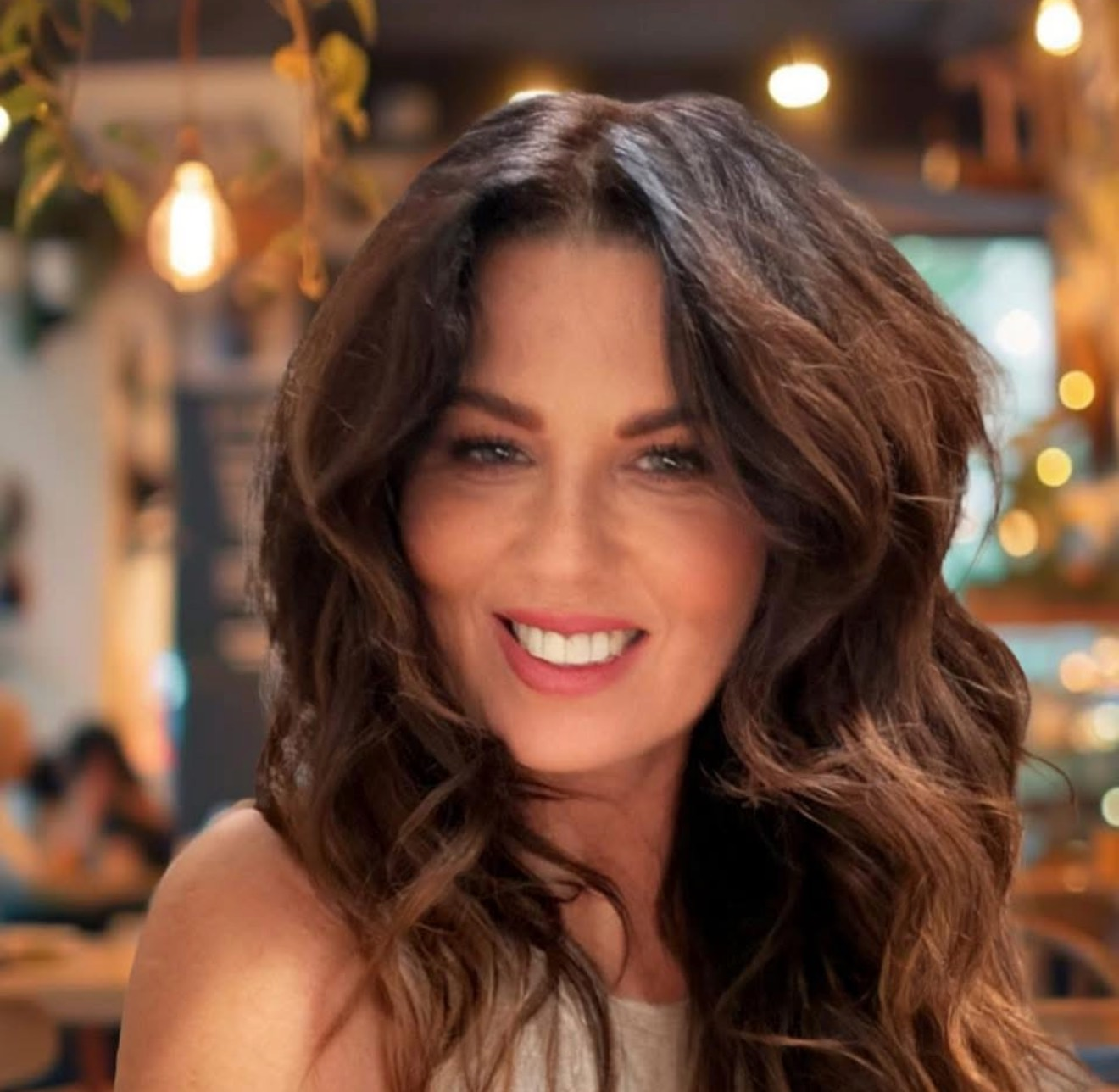 Woman with long, wavy brown hair, smiling with green eyes, wearing a floral top.