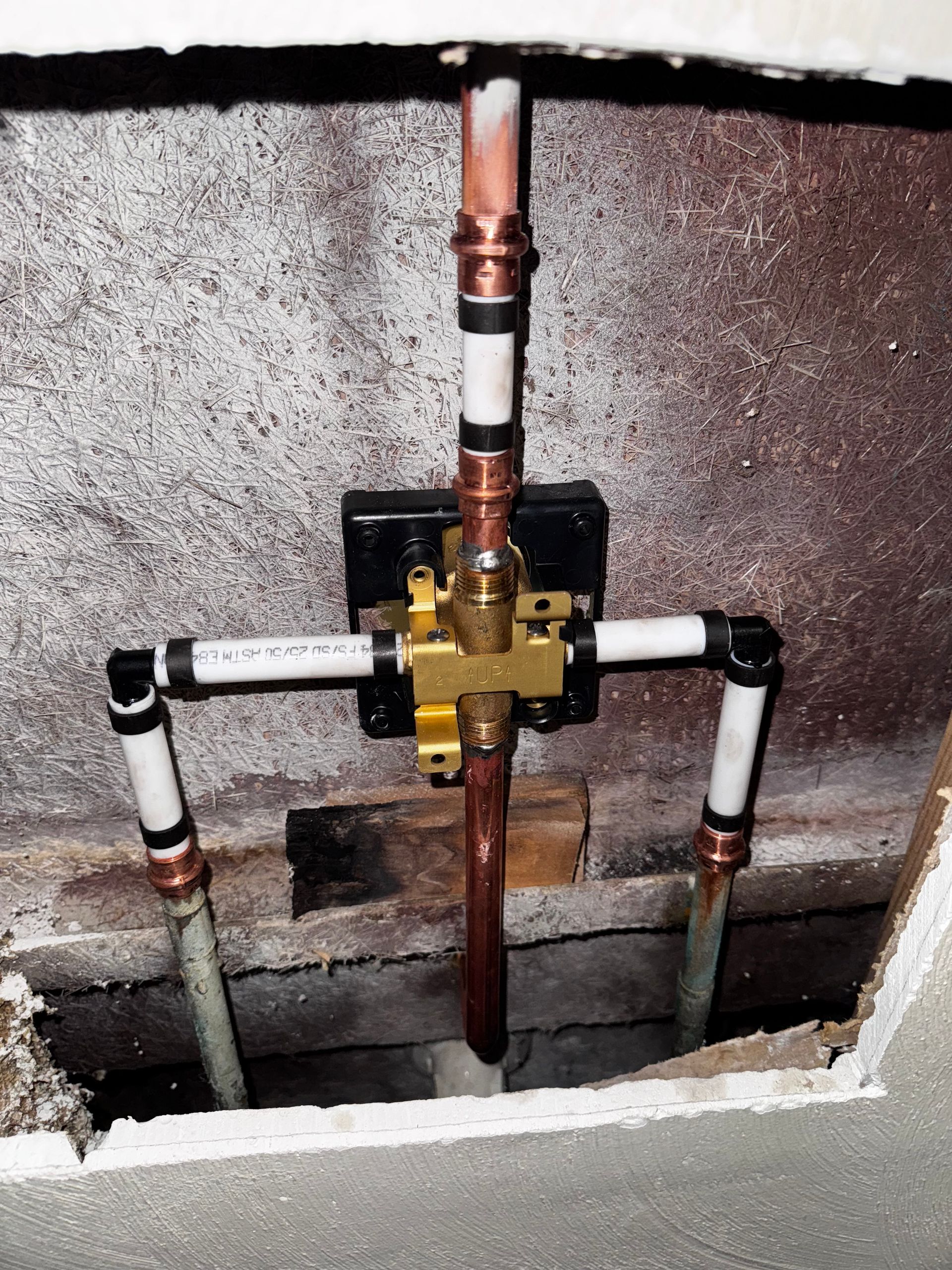 Plumbing with copper and white PVC pipes connected to a brass valve, installed in a wall opening.
