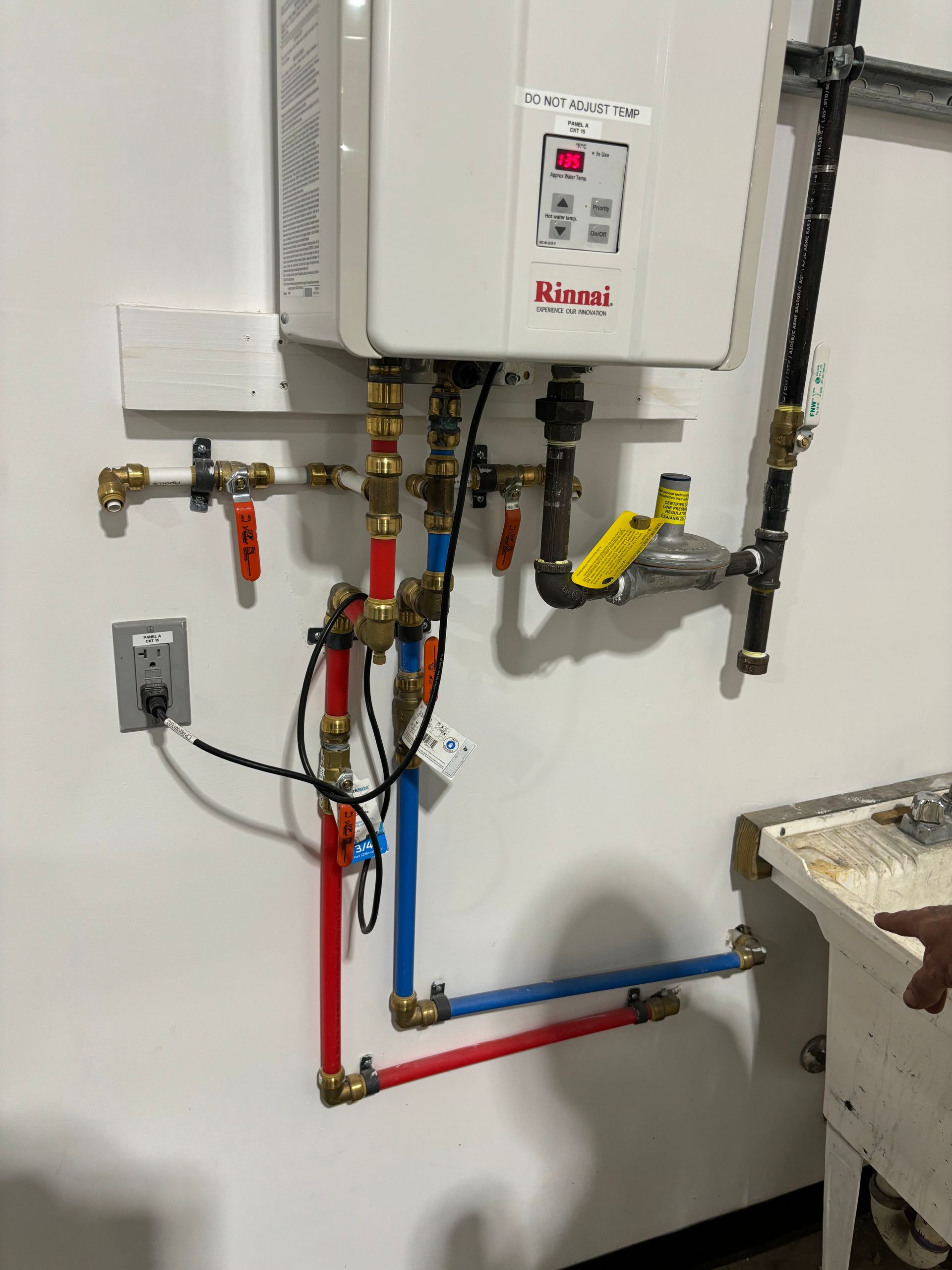 White tankless water heater with connected copper and PEX pipes mounted on a white wall, next to a utility sink.