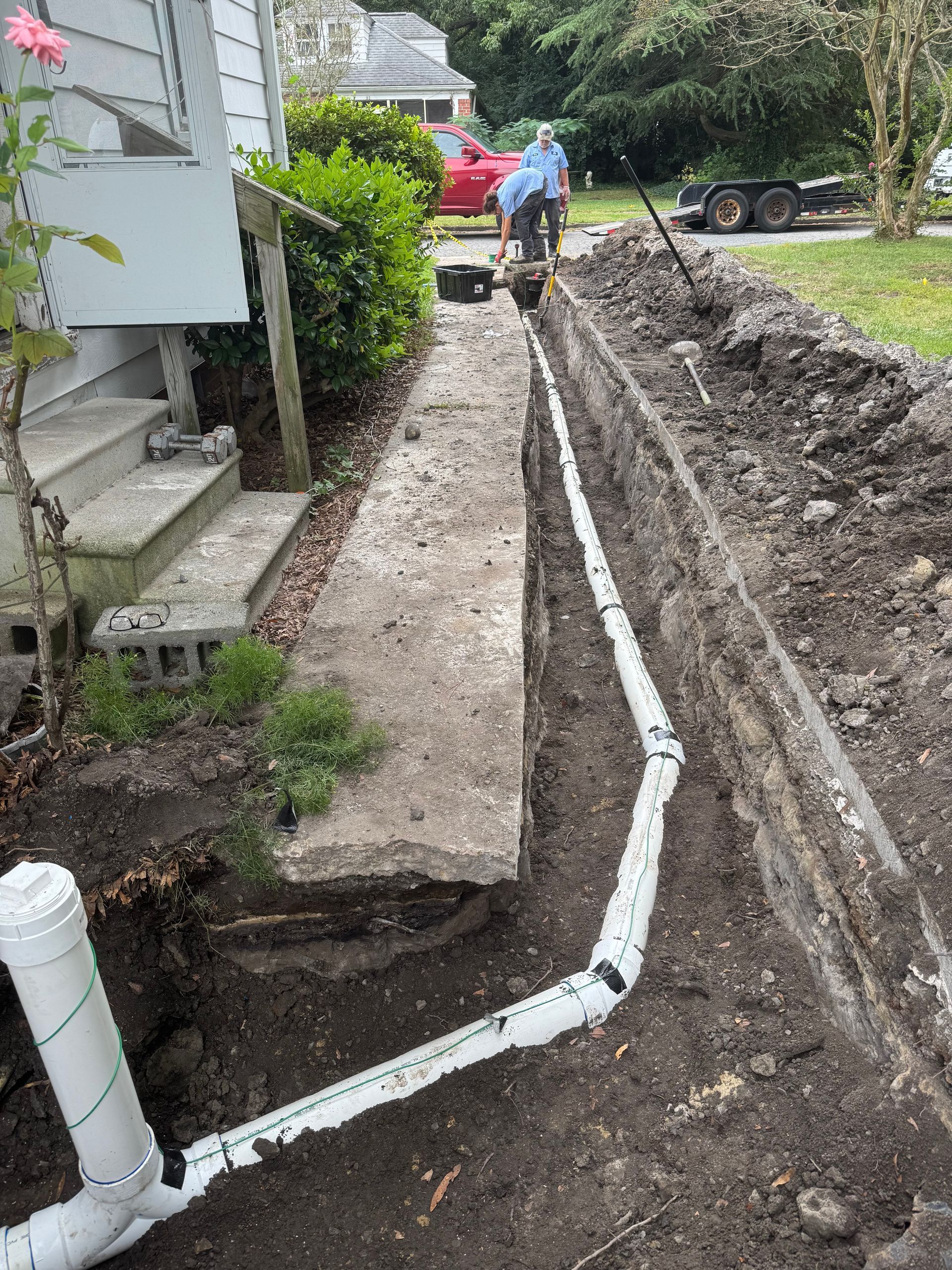 Workers installing white PVC drainage pipes in a trench near a house's foundation.