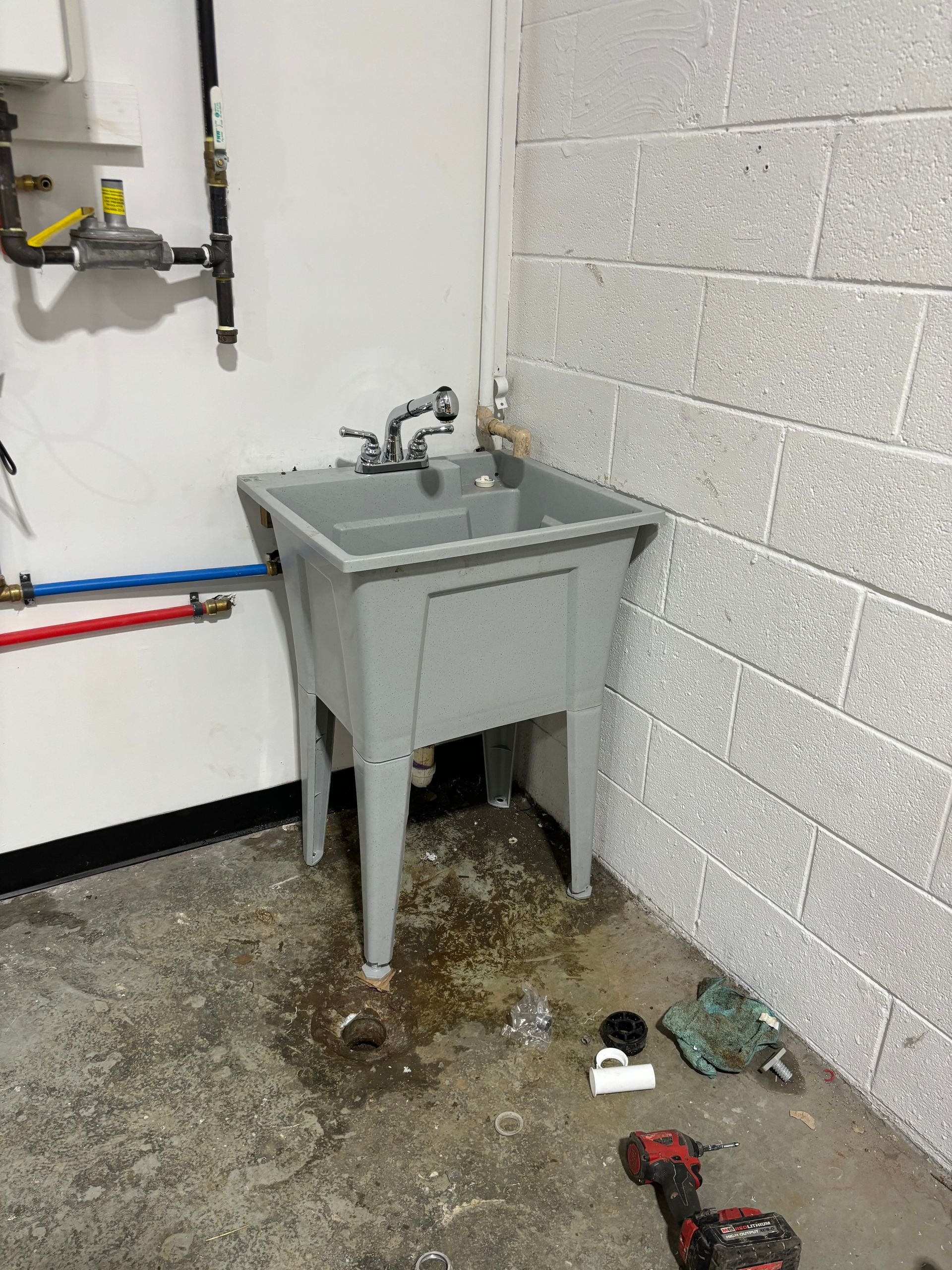 A utility sink in a basement setting, featuring a grey plastic basin and exposed plumbing against a concrete block wall. The floor is stained.