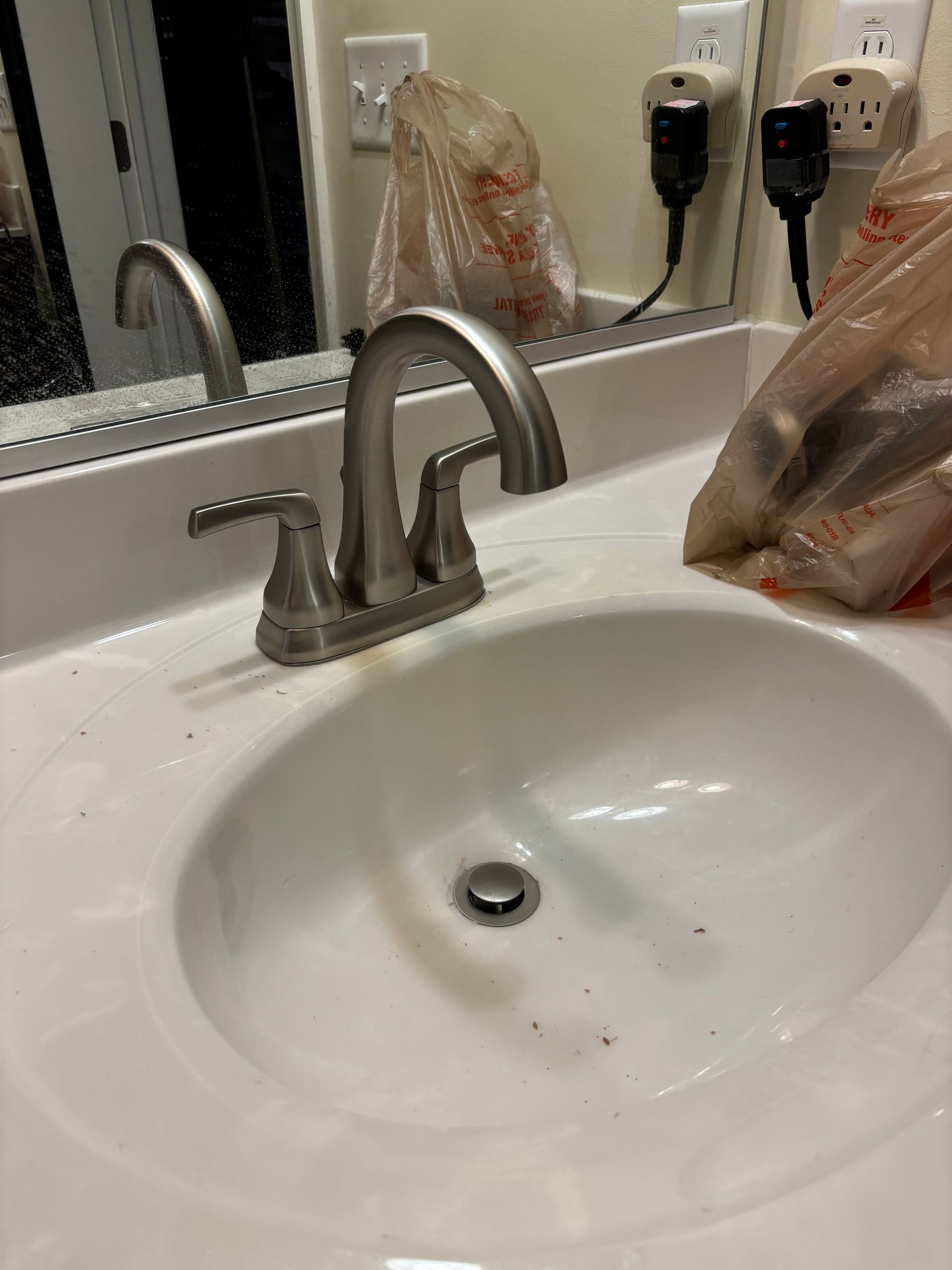 A close-up of a white bathroom sink with a silver faucet, small dark specks, and a bag of items on the side.  The mirror shows a reflection.