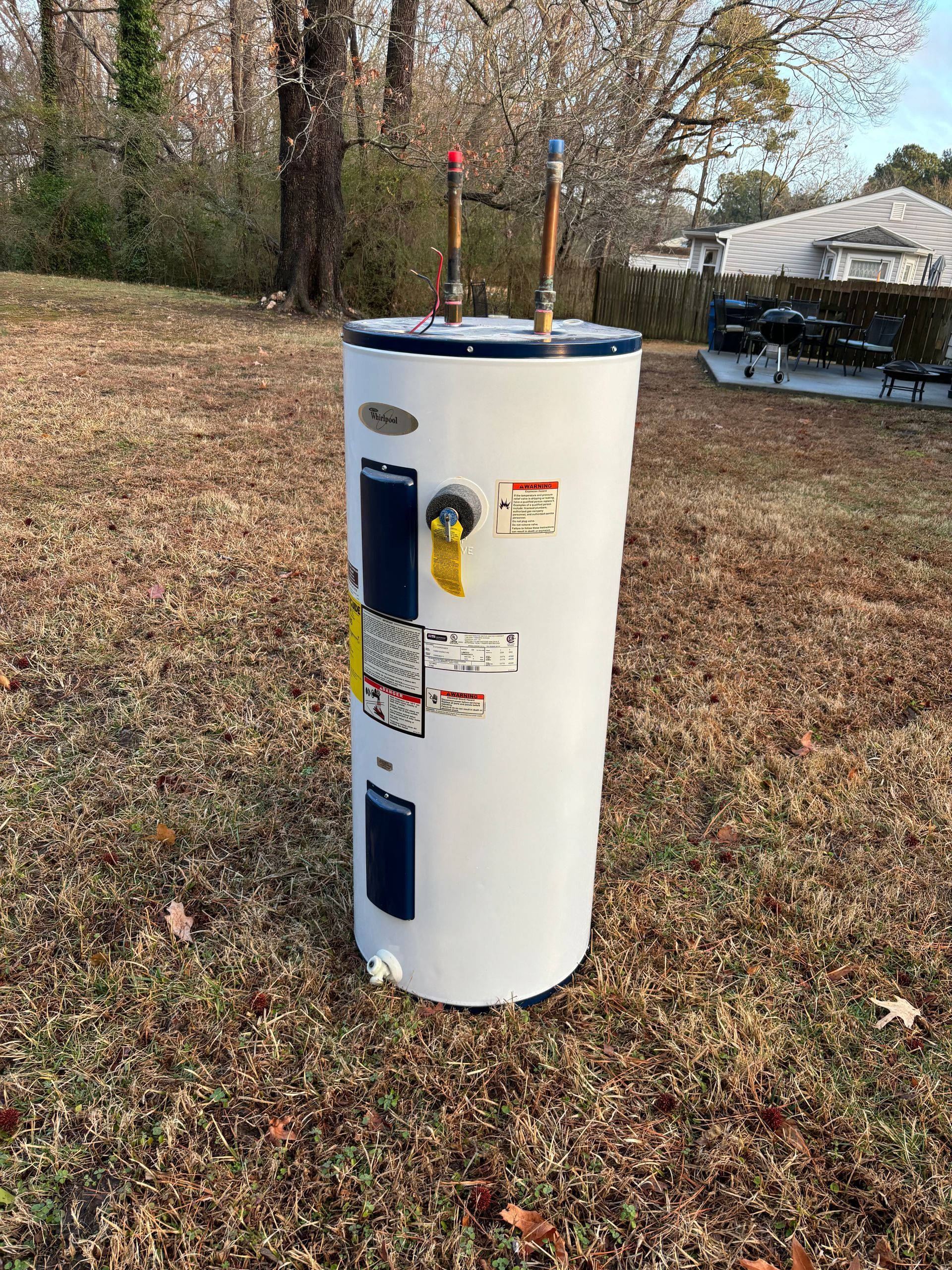 A white Whirlpool water heater stands outdoors in a grassy yard, with copper pipes visible.