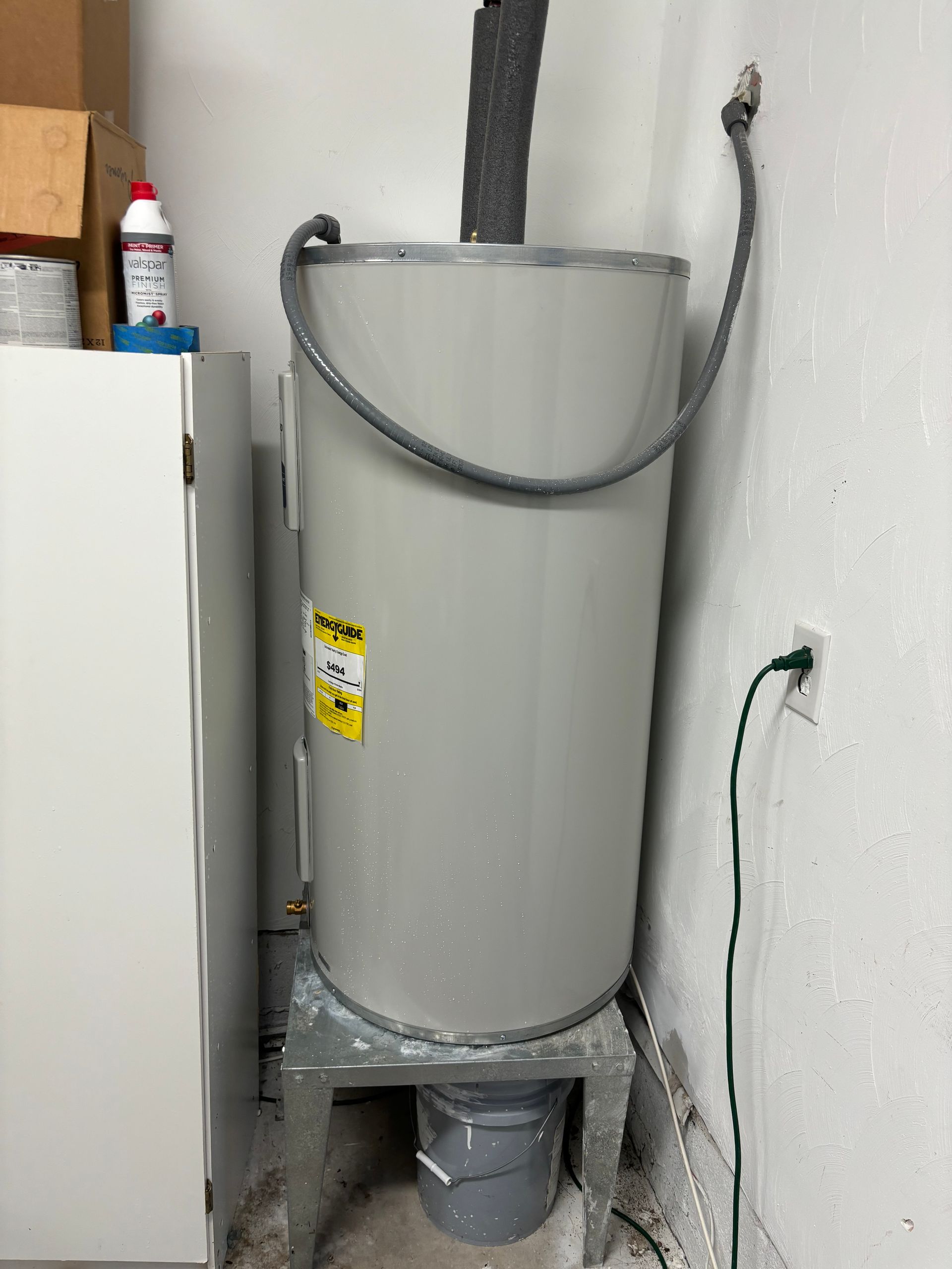 A gray water heater with a black insulated pipe, a gray bucket underneath, and a green power cord plugged into the wall.