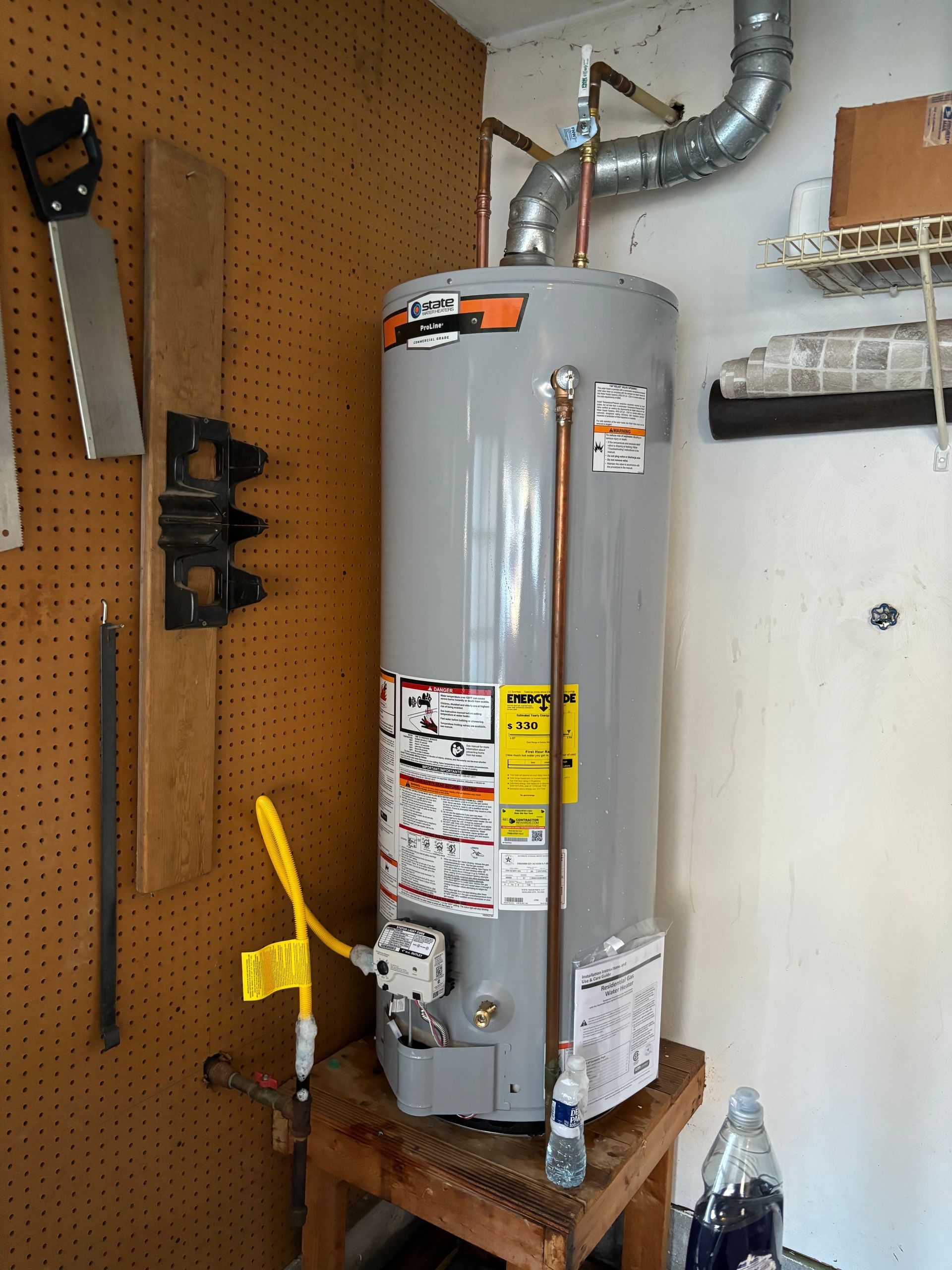 A tall, gray water heater in a garage setting with copper pipes and a vent. Yellow safety tag is visible.