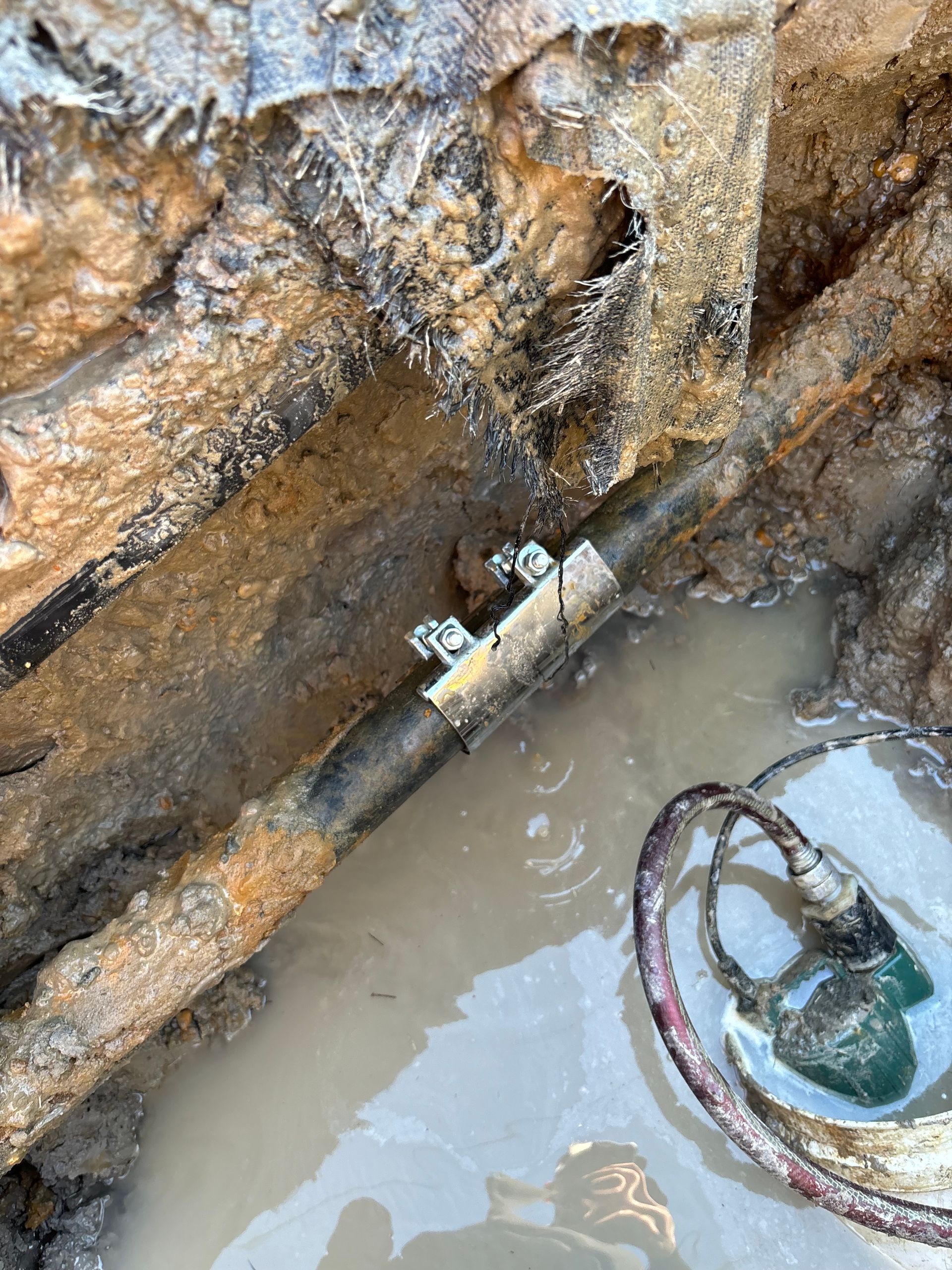 Leaking pipe in a muddy trench, with a metal clamp securing the damaged section. Water pools around the pipe.