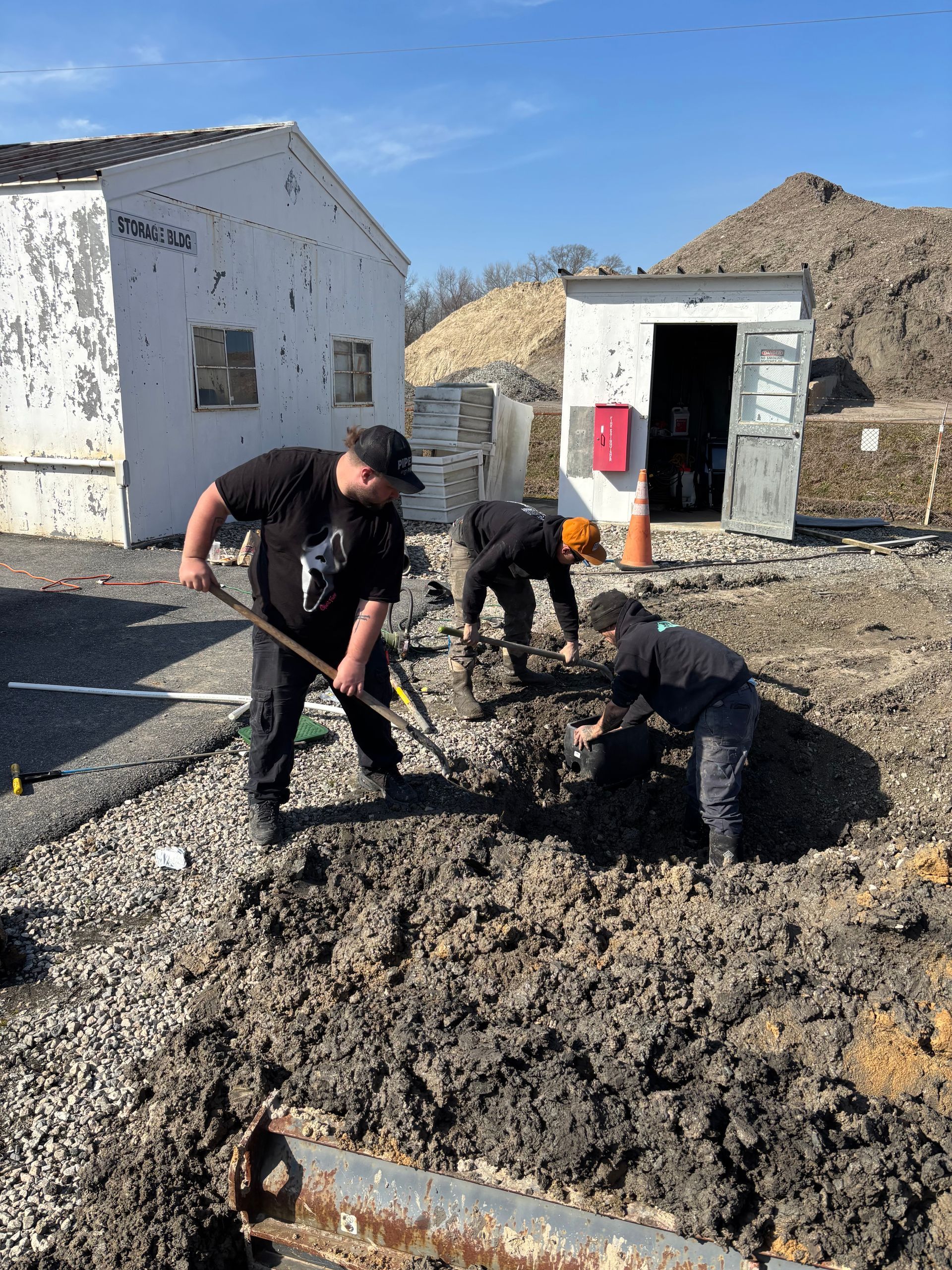 Three people dig in muddy soil with shovels near a white building under a blue sky.