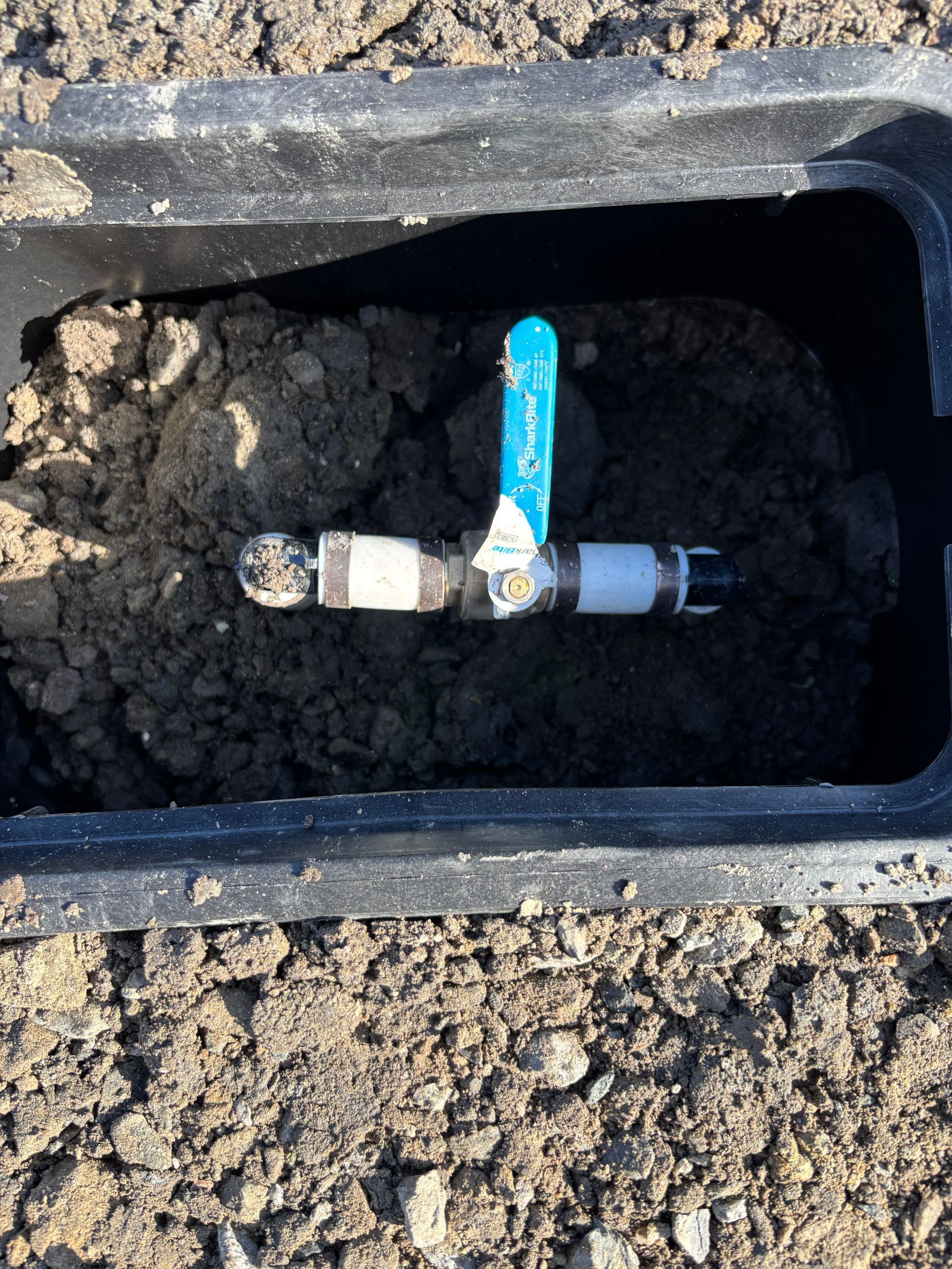 A black irrigation valve box with soil containing a blue-handled valve and connected pipes.