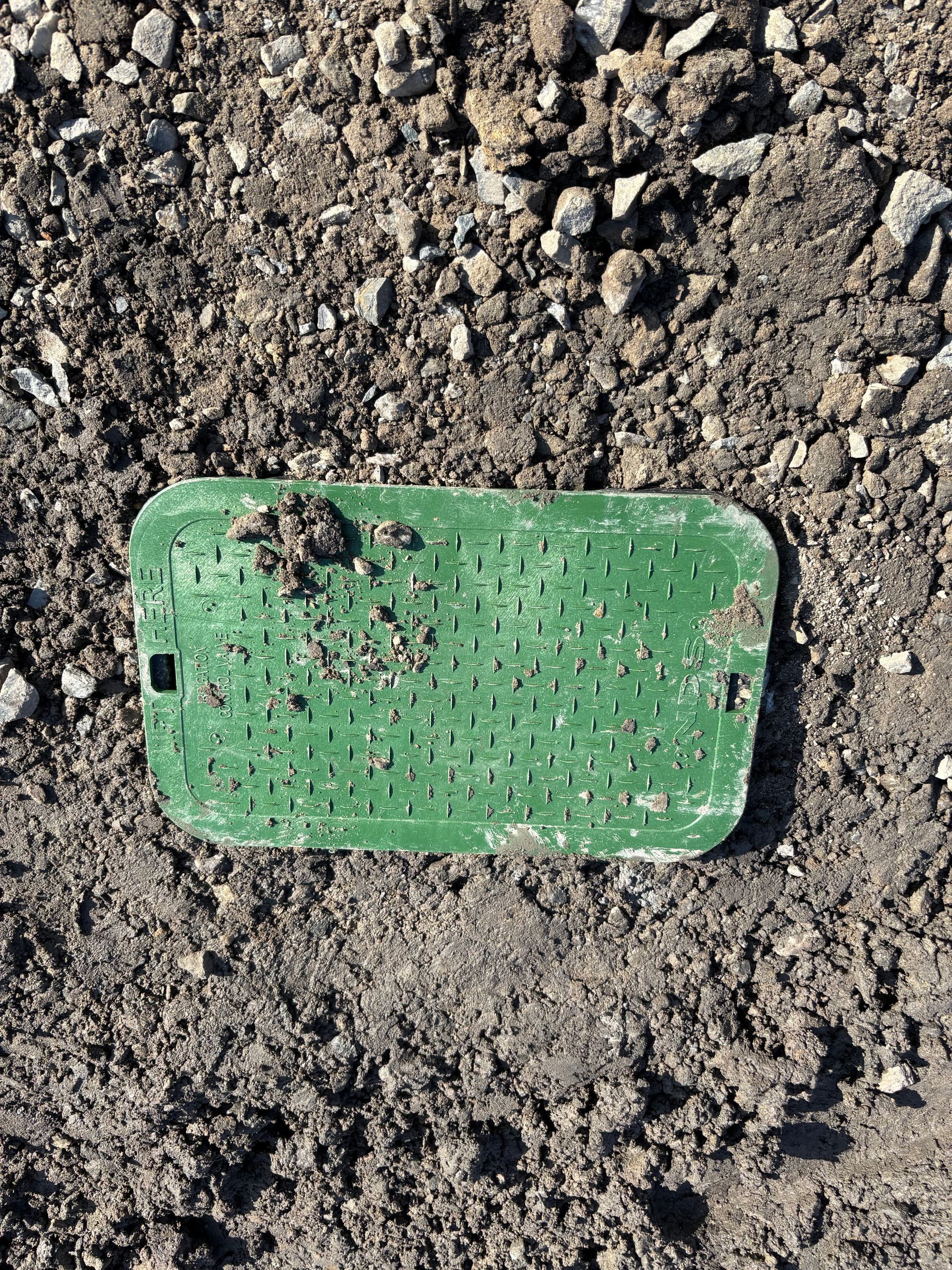 Green rectangular access cover embedded in dirt and gravel.
