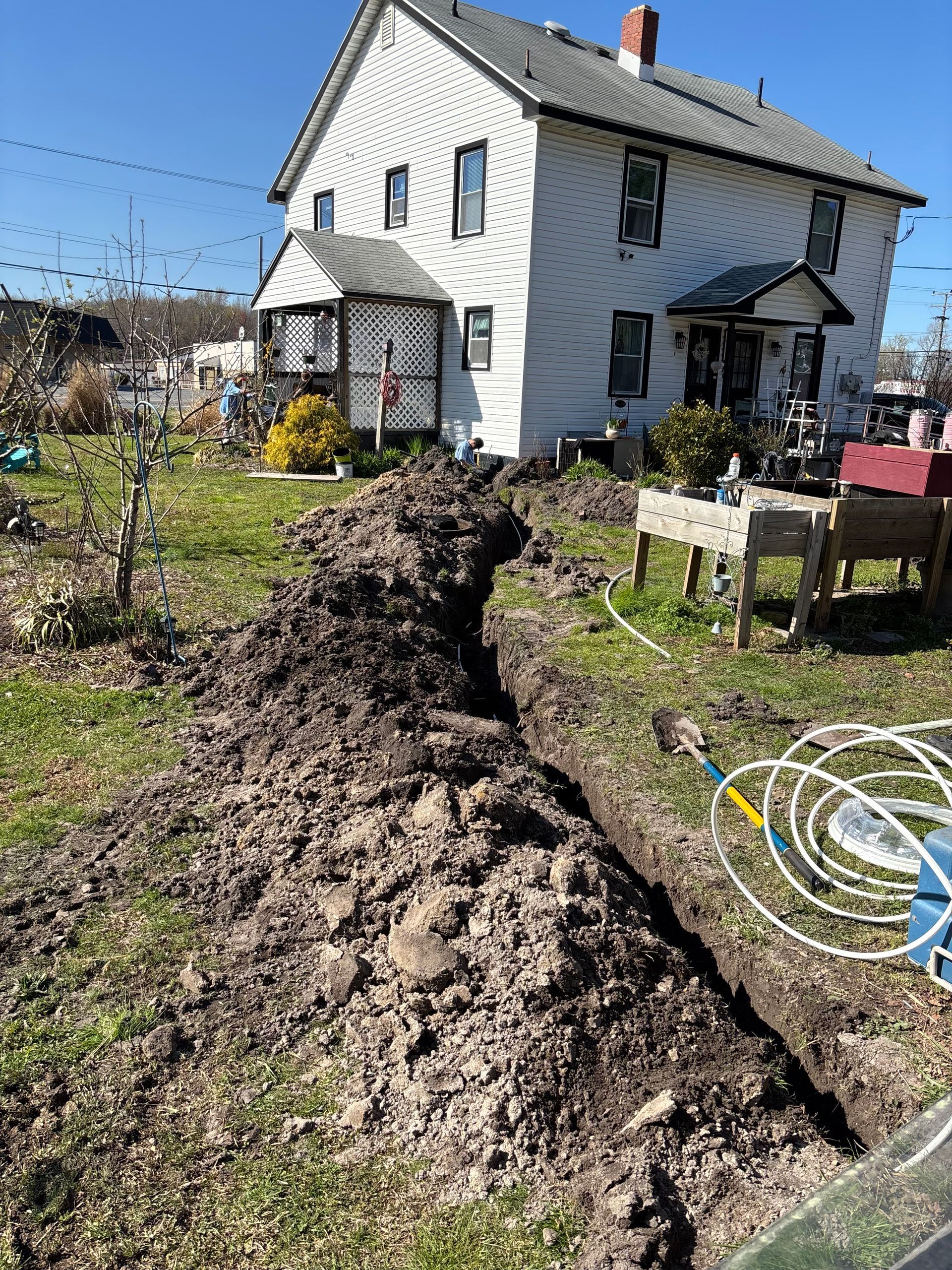 A long trench dug in a grassy yard in front of a two-story white house, likely for utility work.