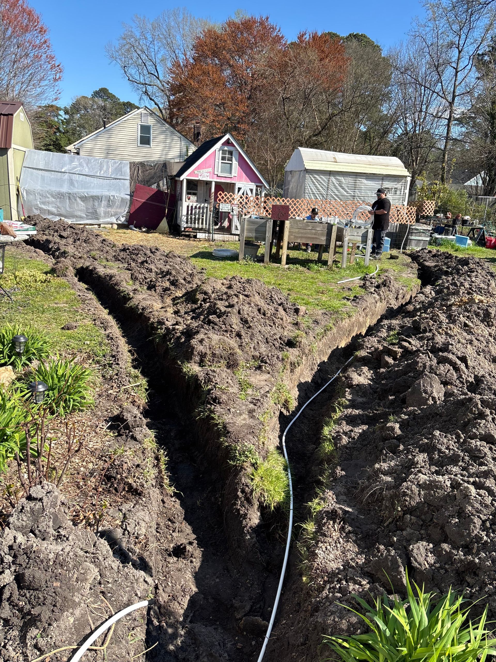 Trenches dug in a backyard garden, with a small pink shed and houses in the background. White electrical cable is visible in the trench.