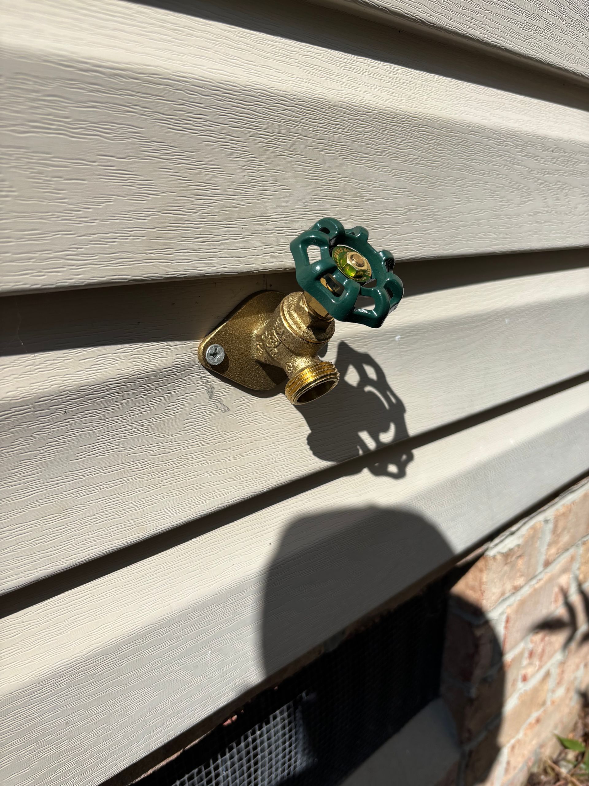 Brass outdoor water faucet with a green handle on beige siding.  Sun shadow on the wall.