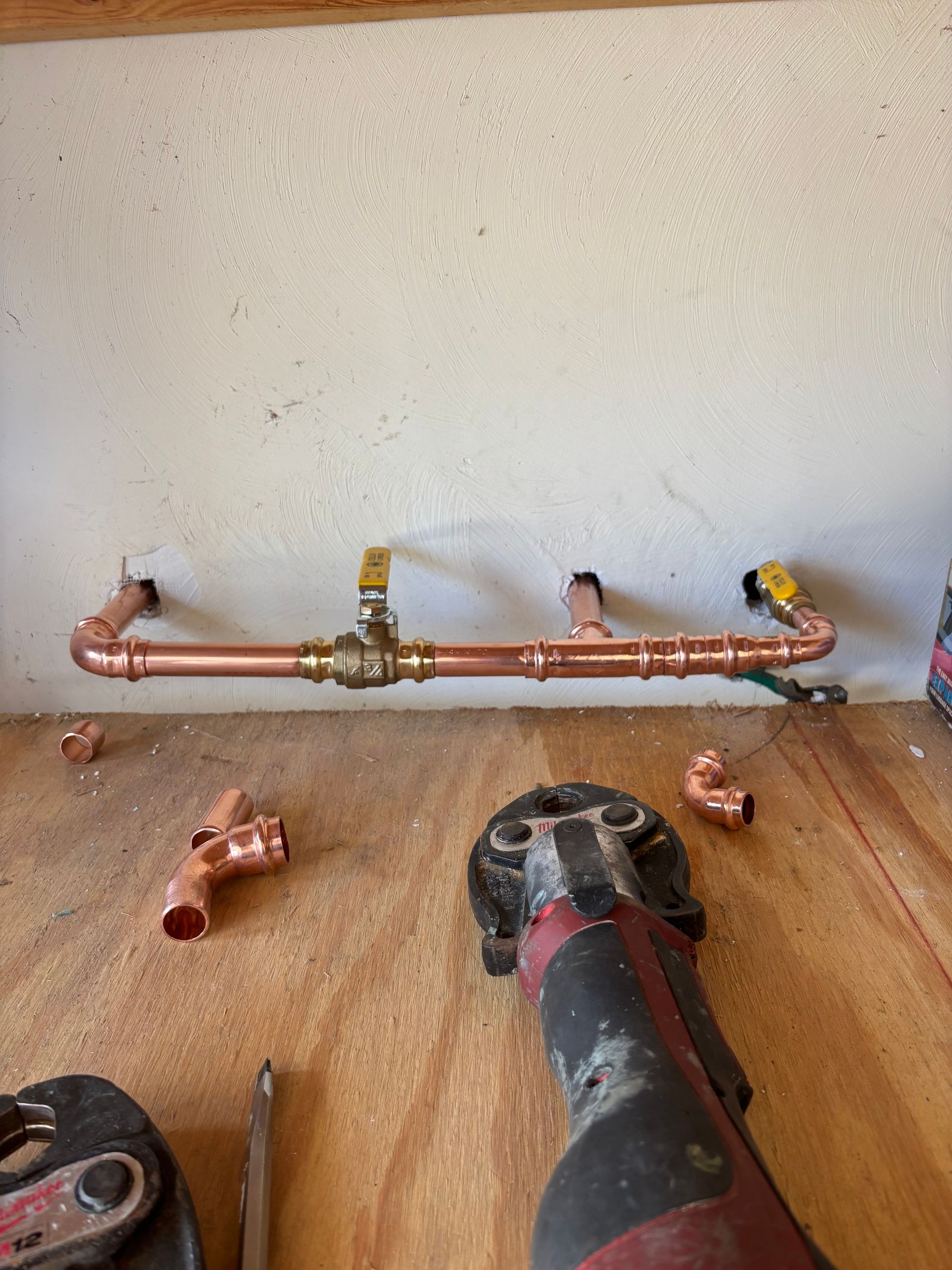 Copper pipes, a valve, and fittings are being assembled with a press tool on a wooden surface, likely for a water system.