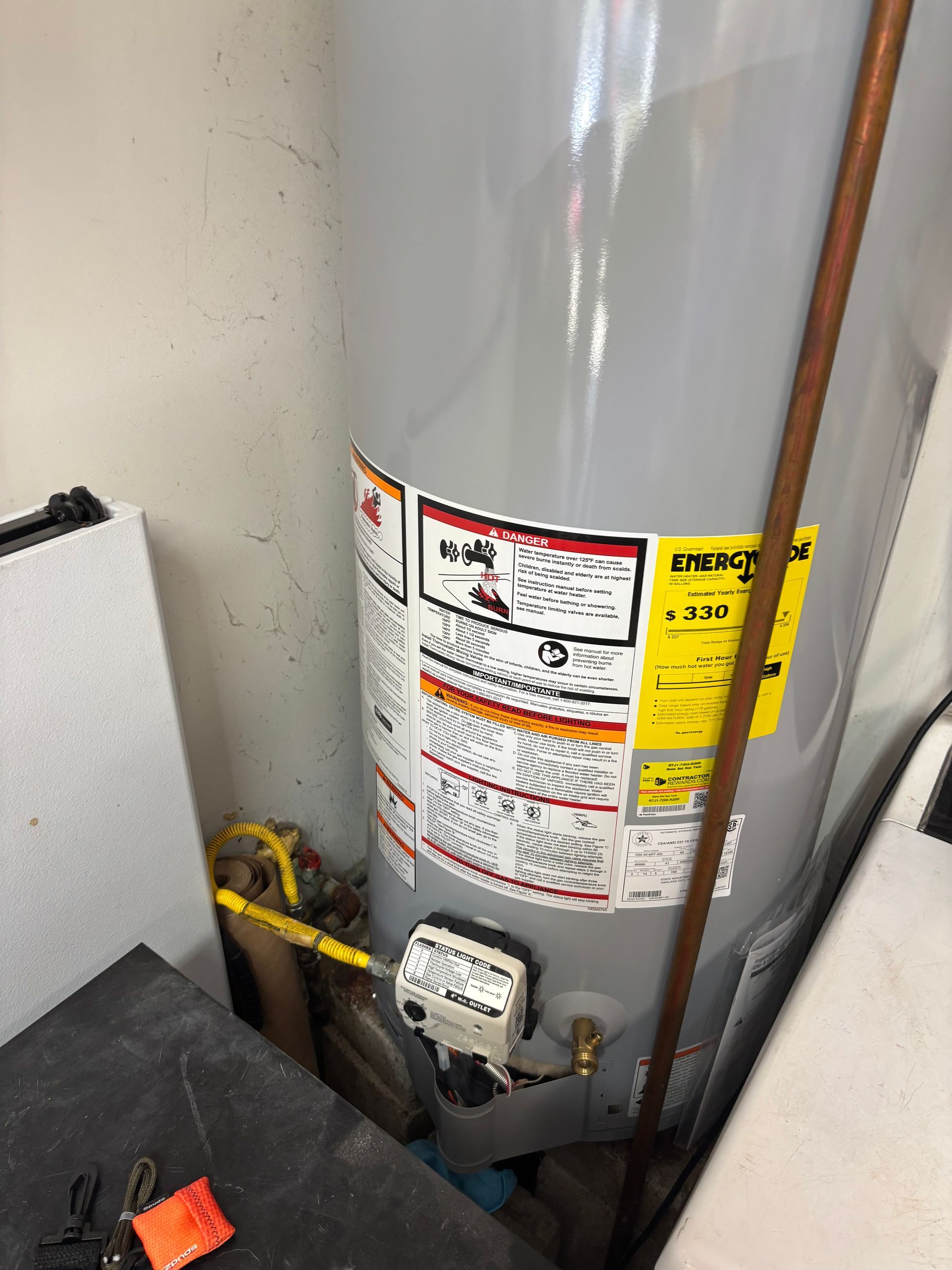A tall, gray water heater in a utility space, with yellow warning labels and copper plumbing. The setting appears to be a garage or basement.