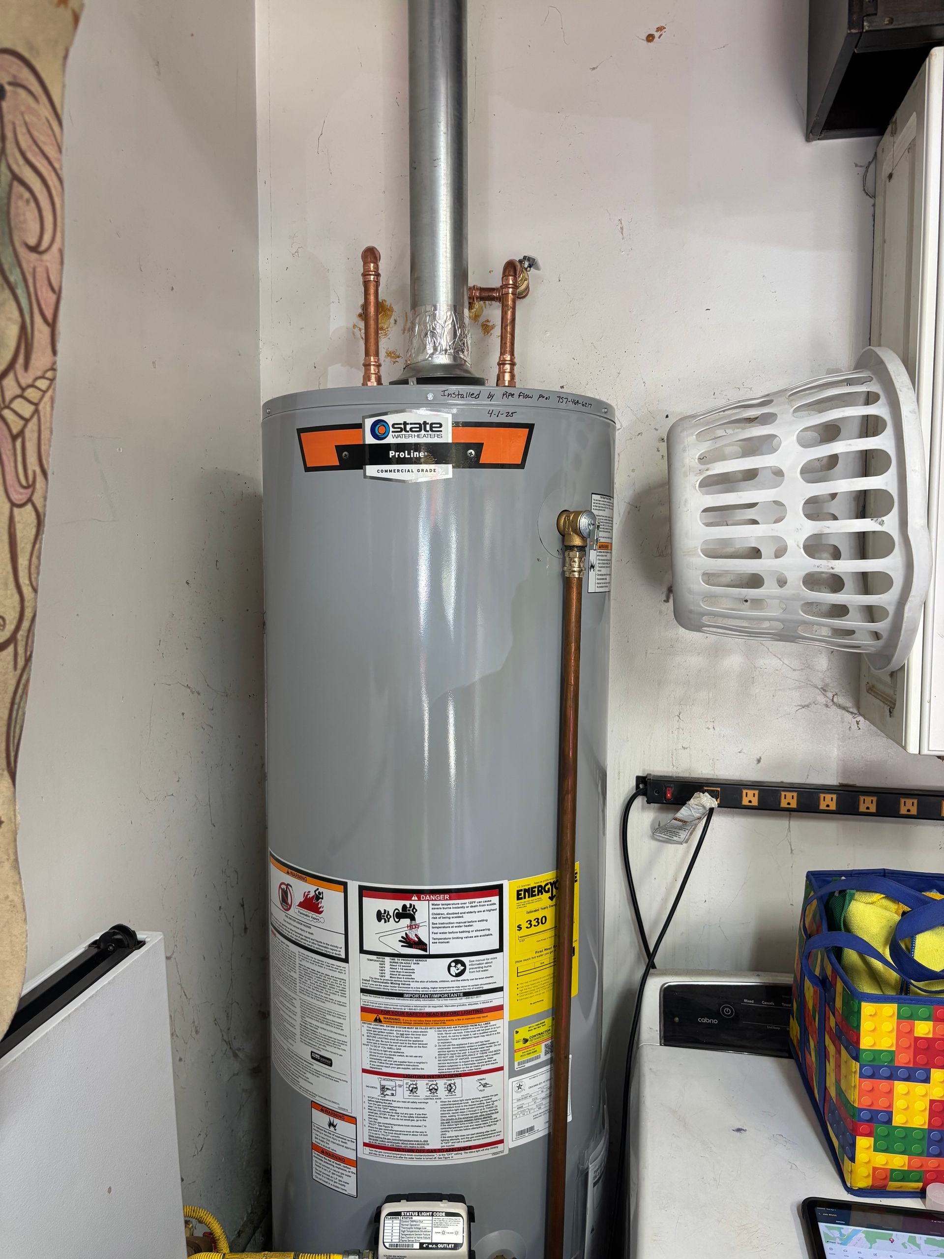 A tall gray water heater in a corner, copper pipes visible, with warning labels, a laundry basket, and a table.