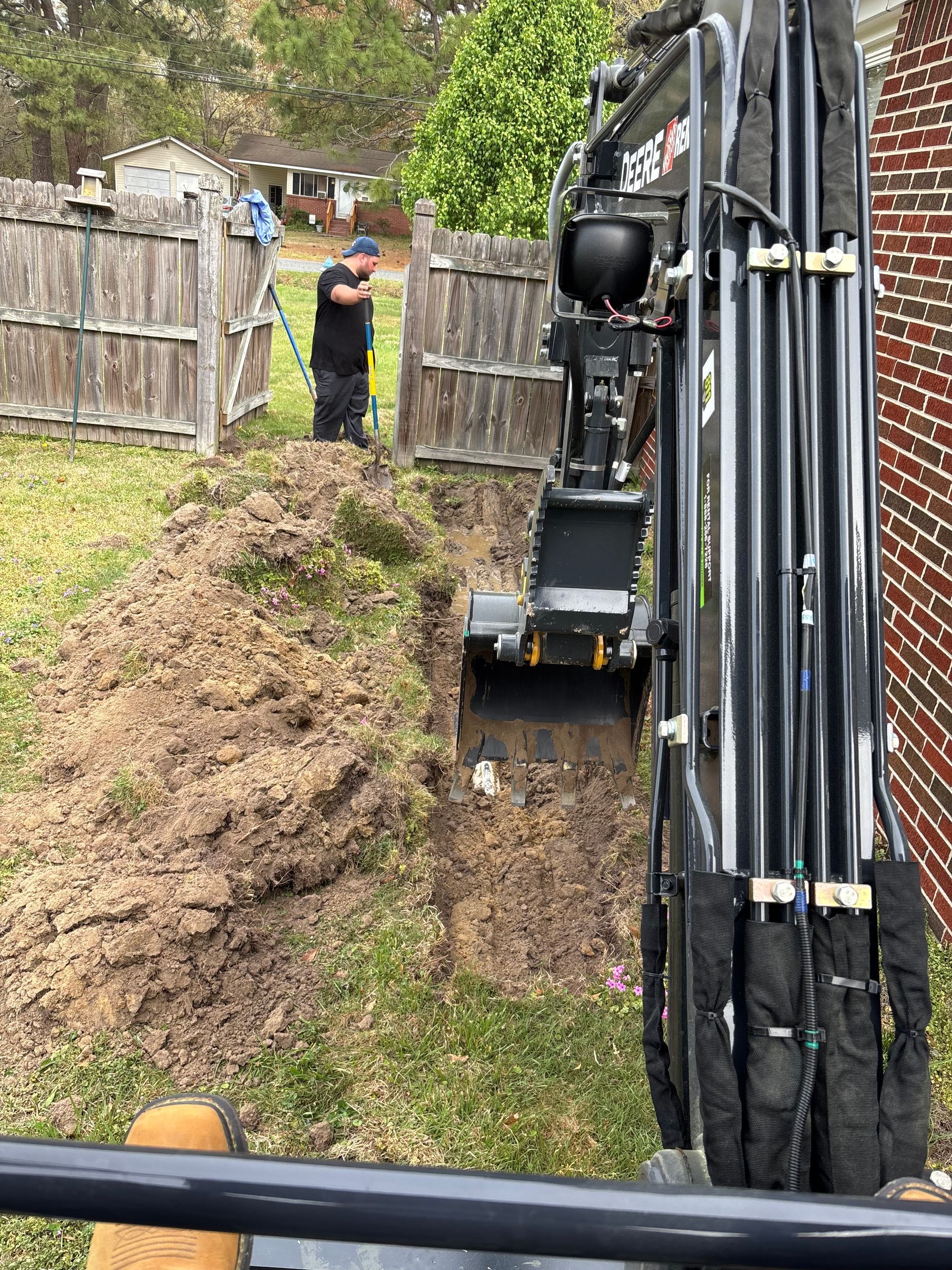 A worker operating a mini-excavator digs a trench in a grassy backyard. A wooden fence and brick building are in the background.