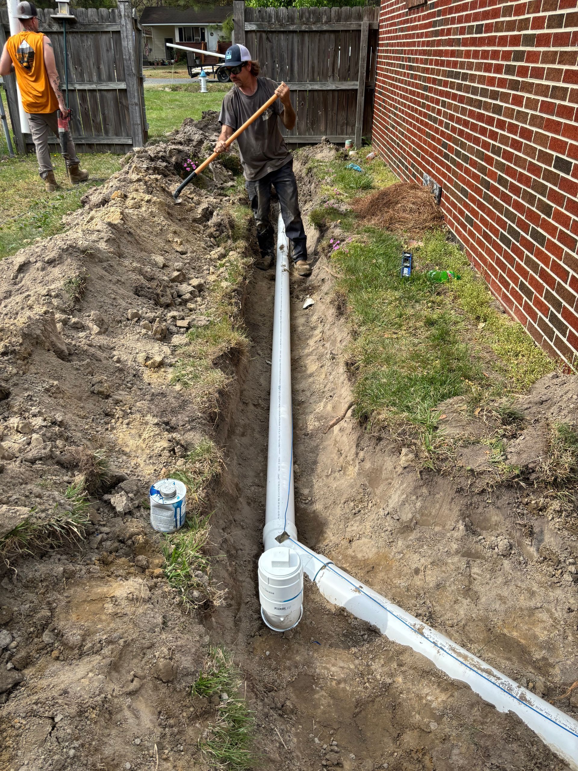 Workers installing drainage pipes in a trench next to a brick building. One worker is using a shovel.