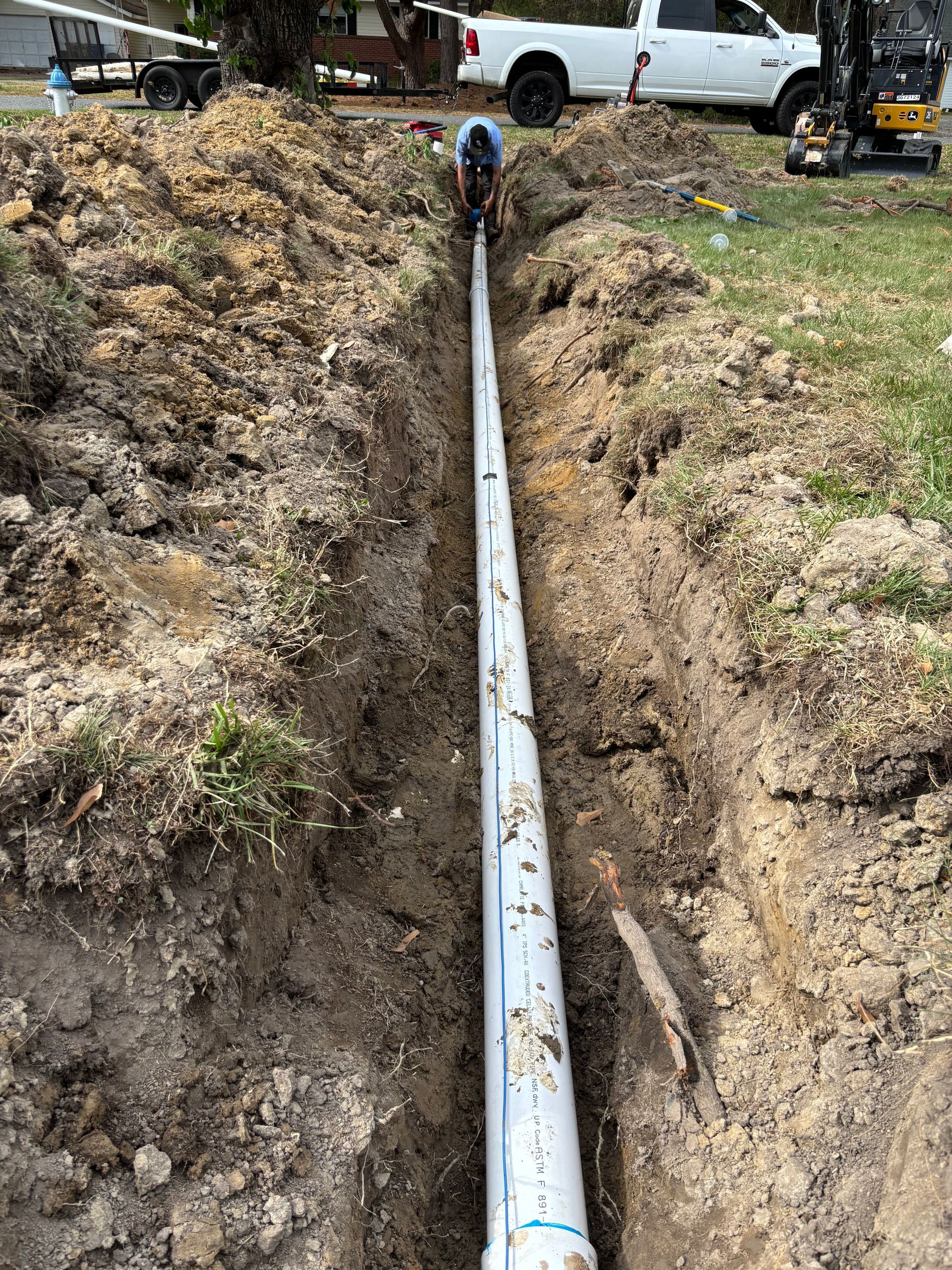 A worker is laying a long white pipe in a narrow trench dug in the ground. A white truck is visible in the background.