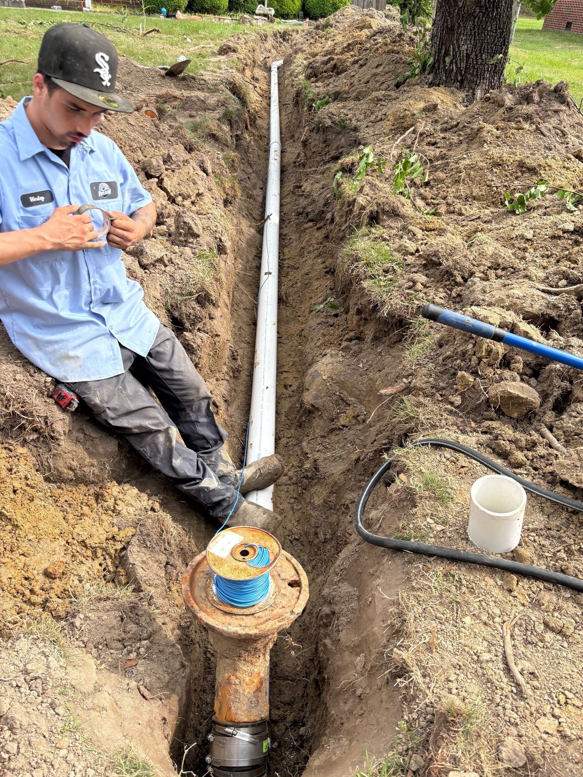 Man installing pipe in a trench. He's wearing blue work clothes and sitting in the trench. A spool of blue wire and other plumbing parts are visible.