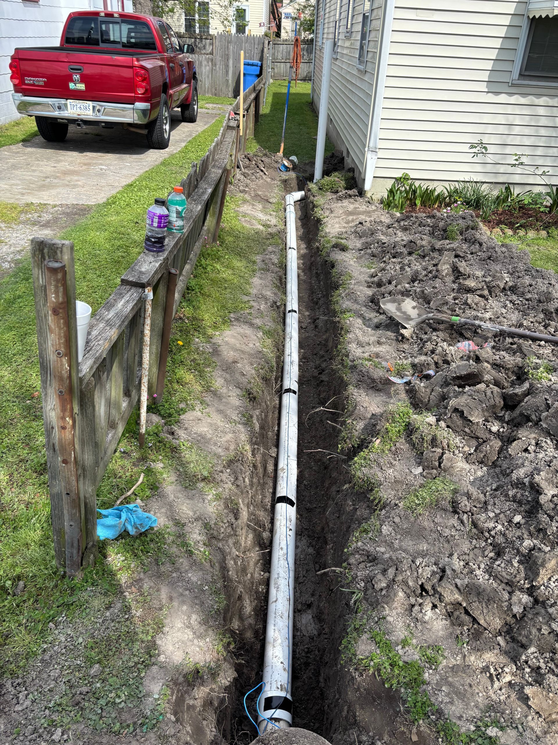 A trench dug in a yard with a white pipe installed, beside a fence and a house. A red truck is parked nearby.