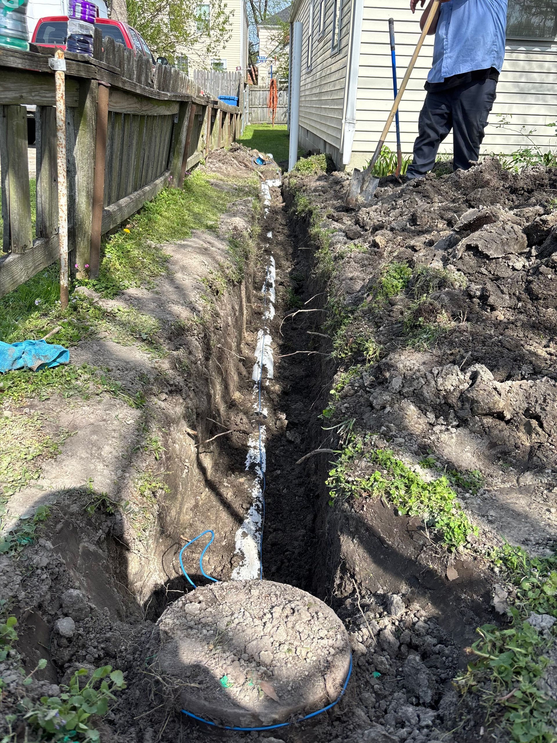 A person digs a trench in a yard next to a house and a fence, with exposed white pipes visible.