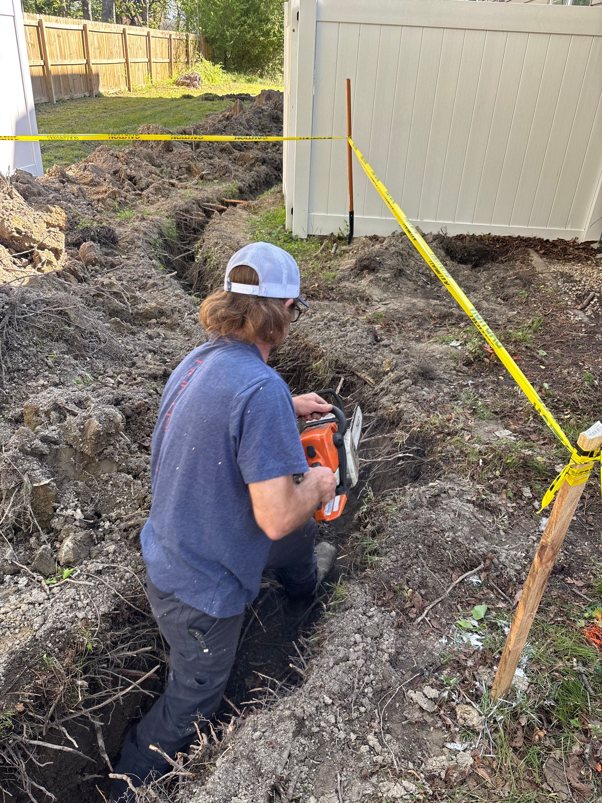 Man using a saw to dig a trench next to a white fence; yellow caution tape overhead.