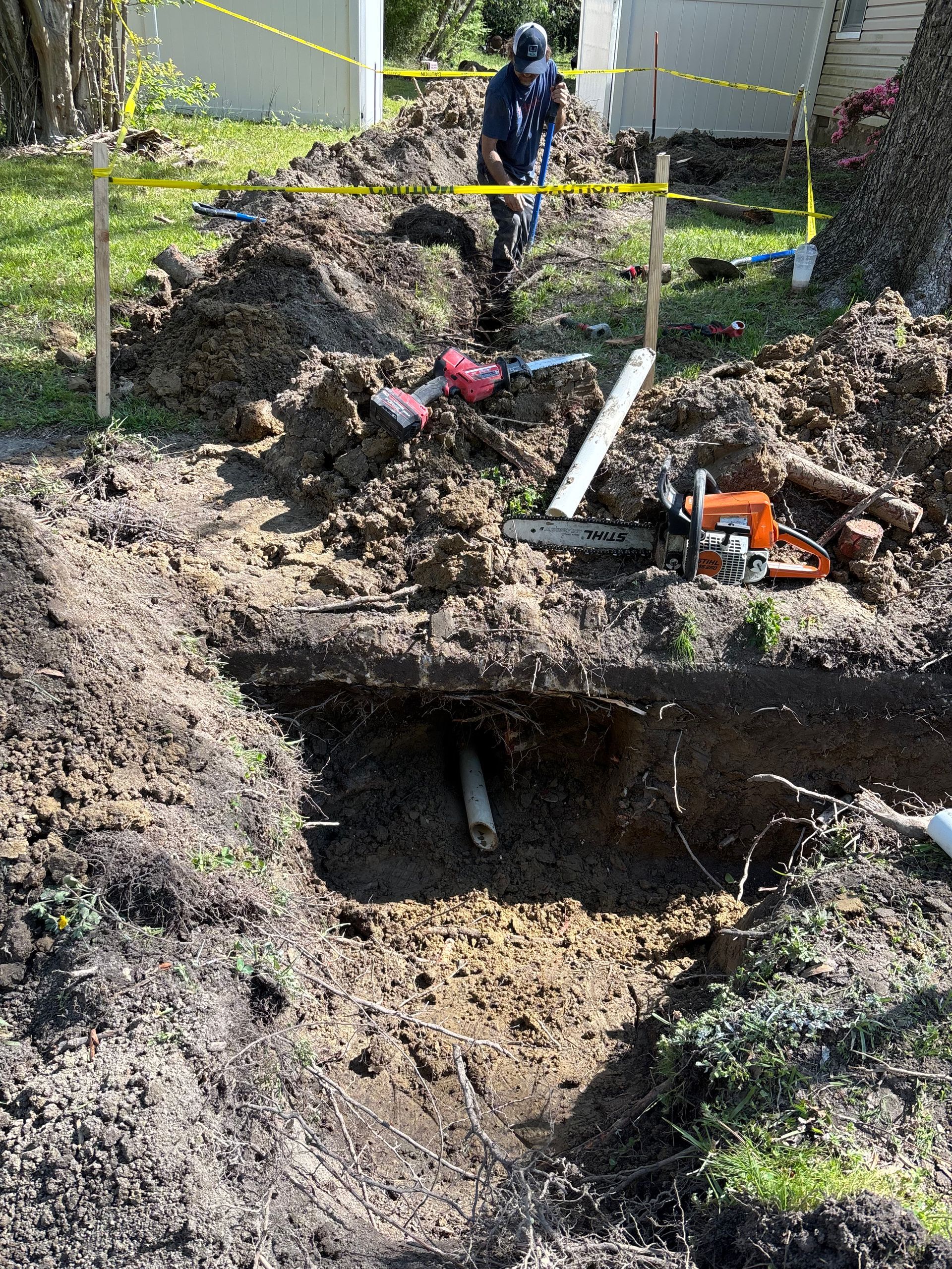 A person digging in a trench outside near a house, with a chainsaw on the edge and a pipe exposed.