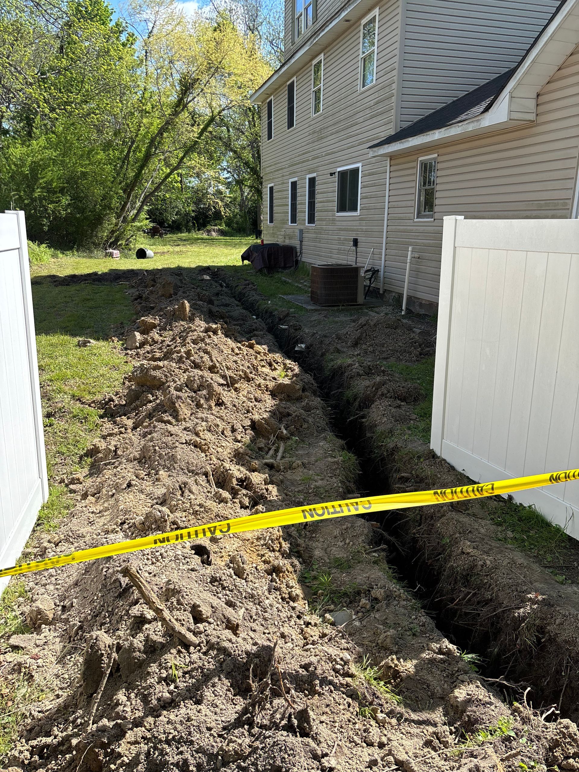 A trench dug in a grassy backyard near a two-story house, blocked by yellow caution tape.  White fence borders the yard.