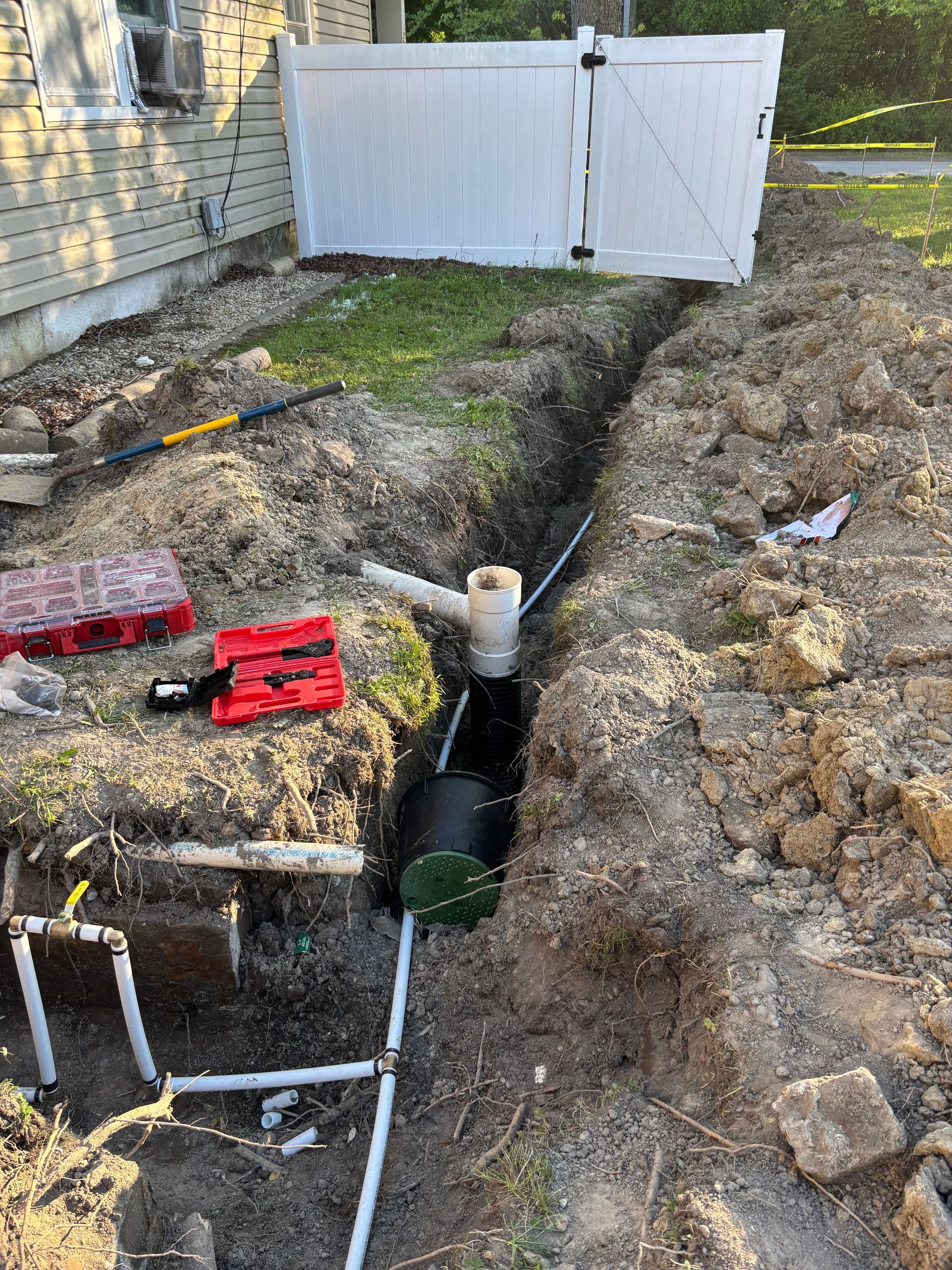 A trench dug in the ground with piping, a septic tank, and tools, near a white fence and a house.