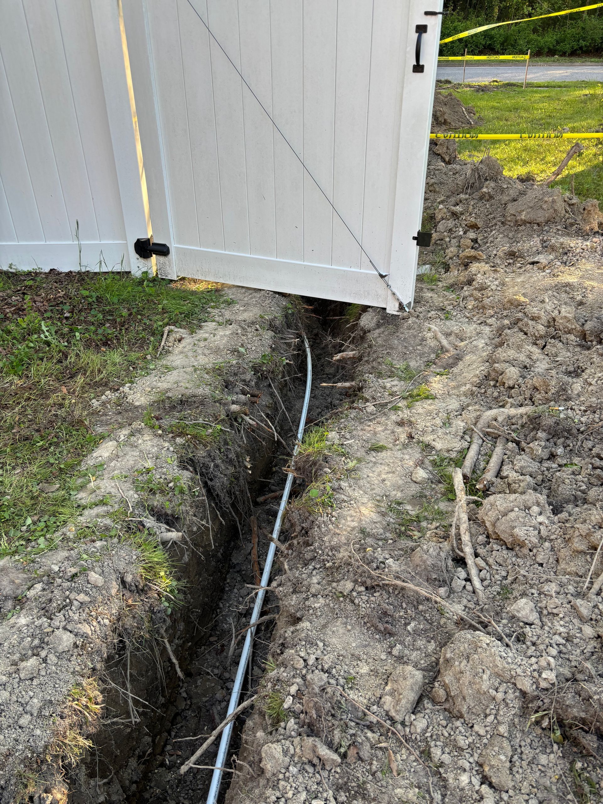 Trench dug near a white fence, containing a silver conduit. Dirt surrounds the trench, in a yard with grass.