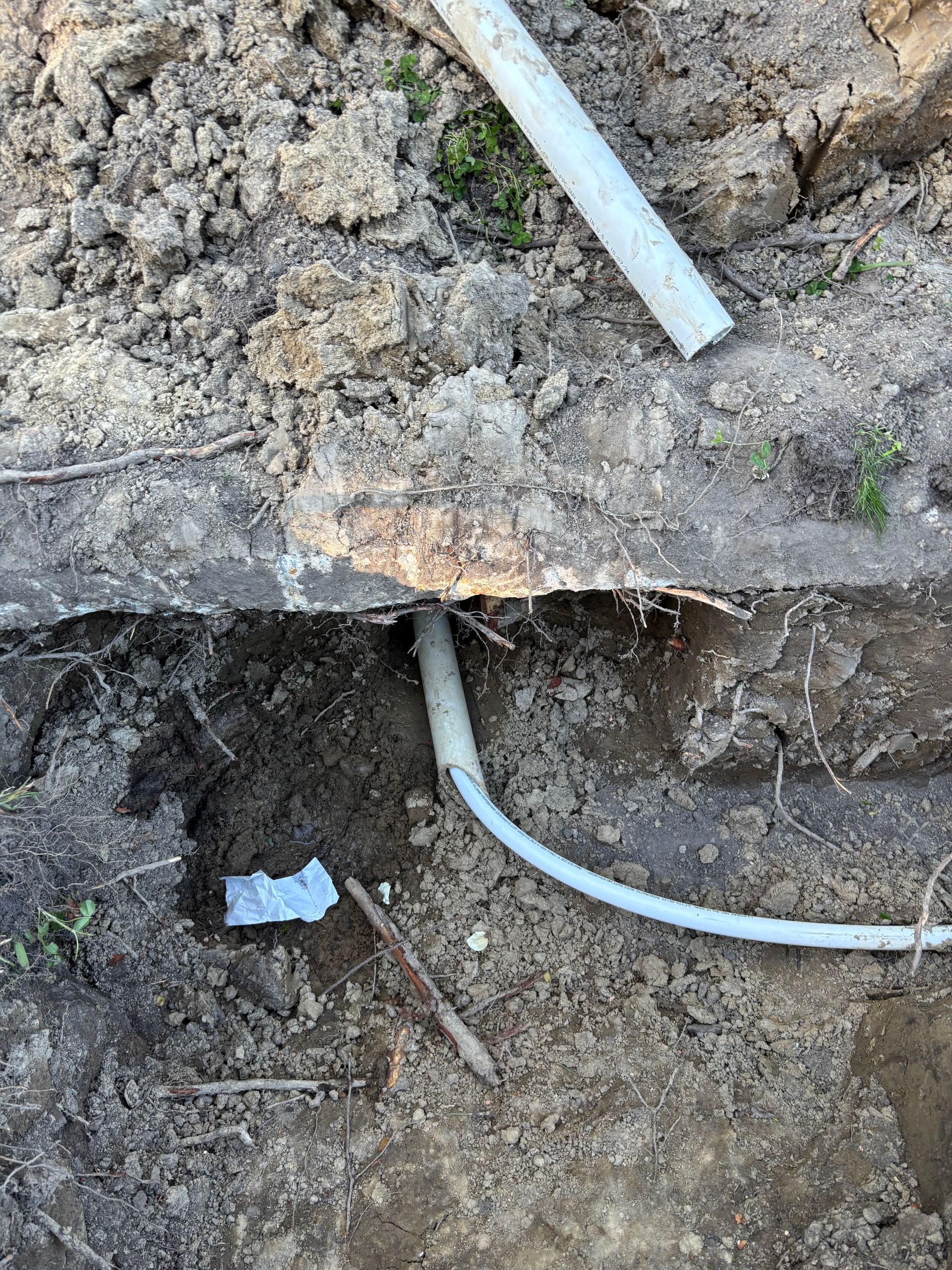 A trench in dirt reveals a white pipe entering a concrete slab, with another pipe section above and debris nearby.