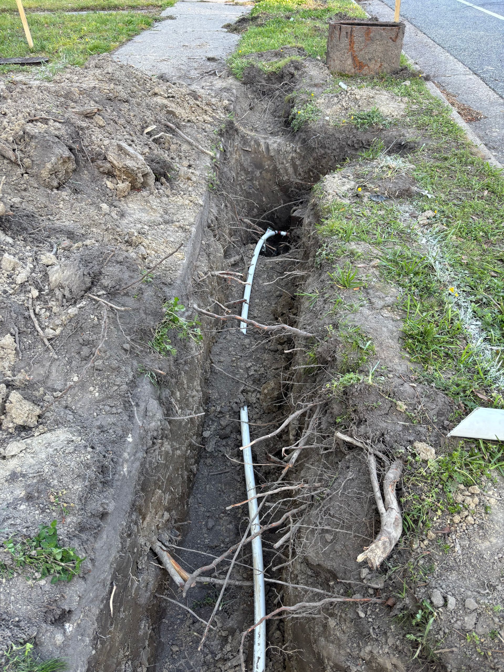 Trench dug alongside a road, exposing white pipes and tree roots. A shovel rests on the edge.