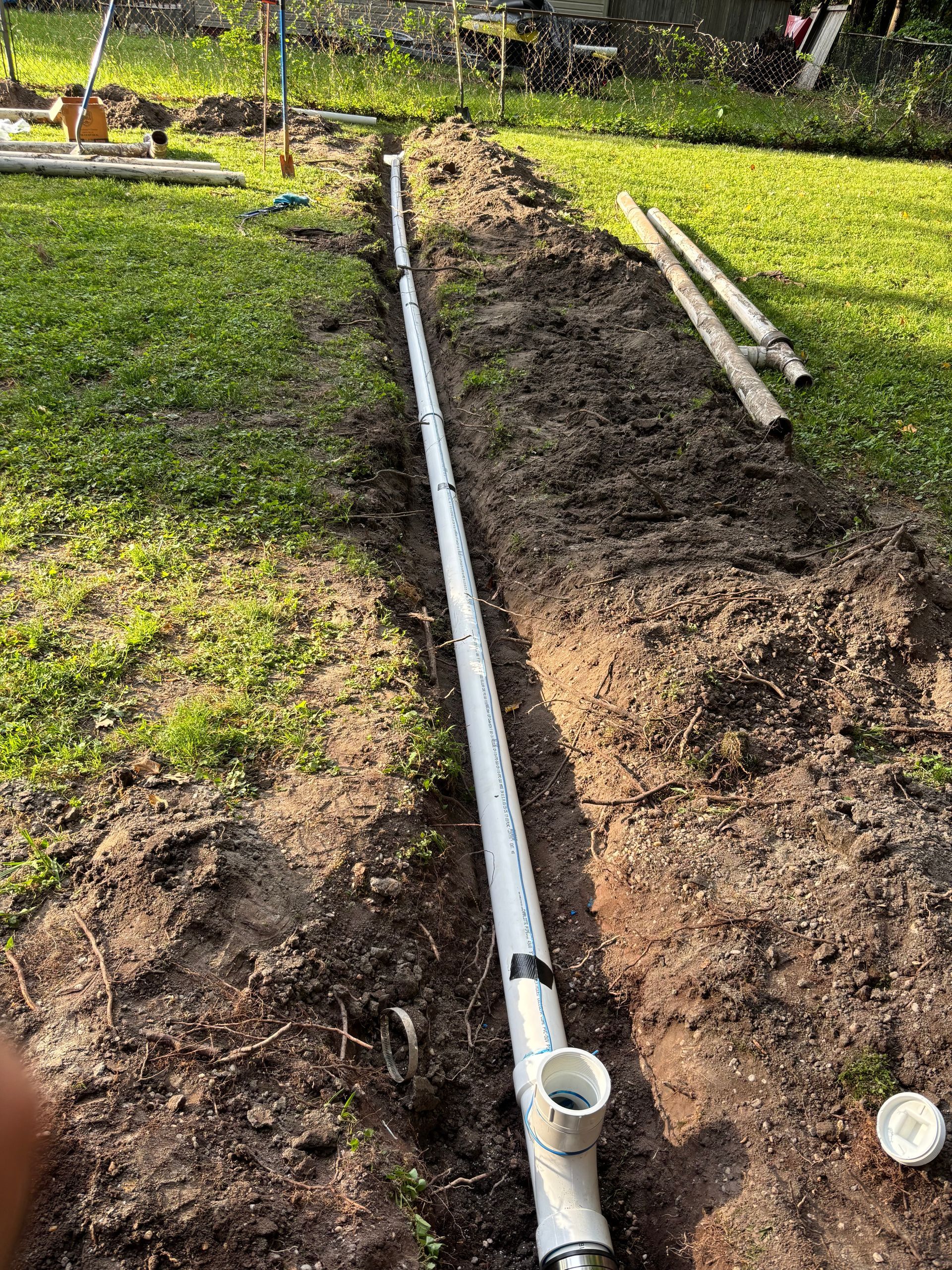 A long trench in a yard with white PVC pipe laid inside, likely for drainage, along with some dirt and tools.