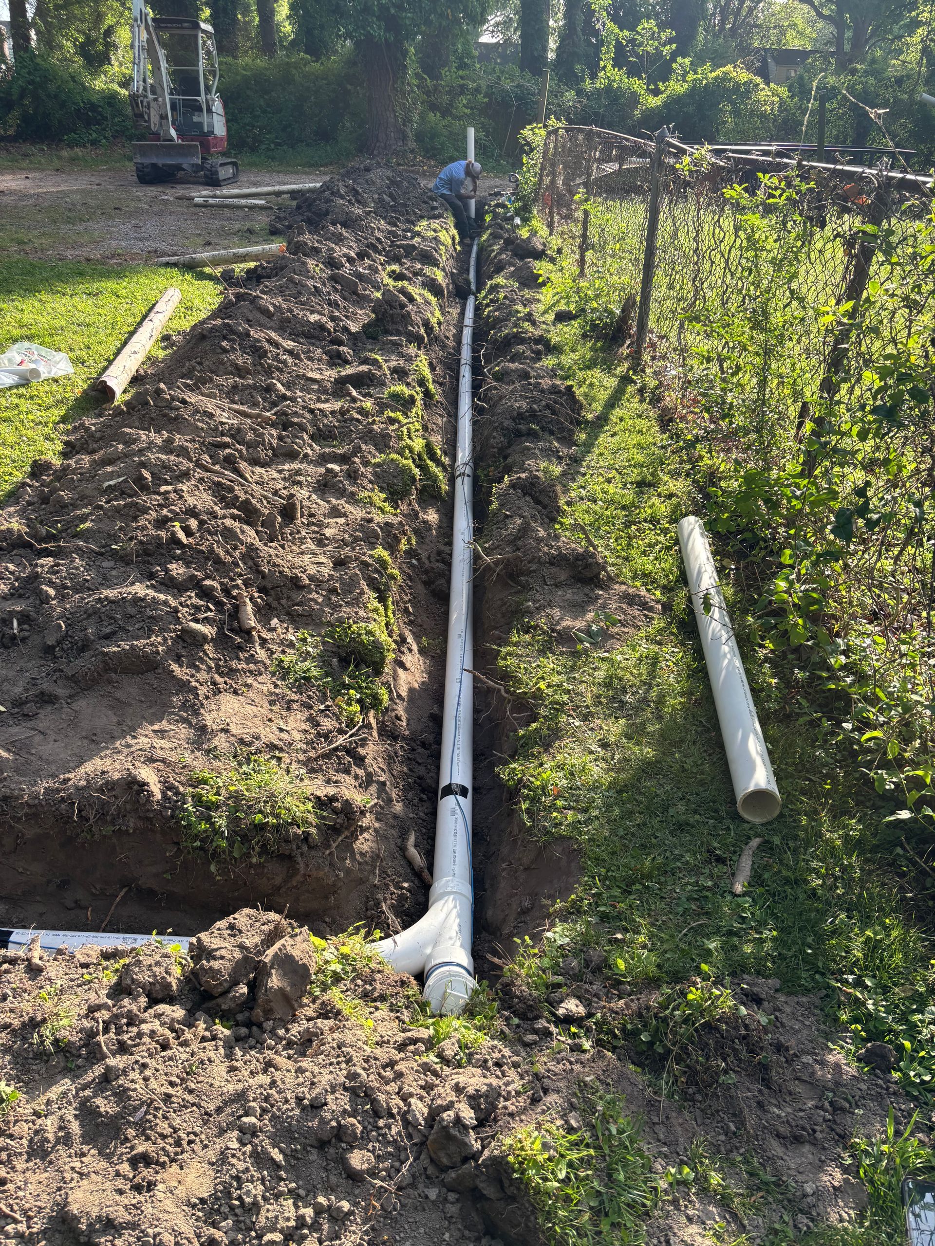 A trench dug in a yard with white PVC pipes running through it, presumably for plumbing or drainage.