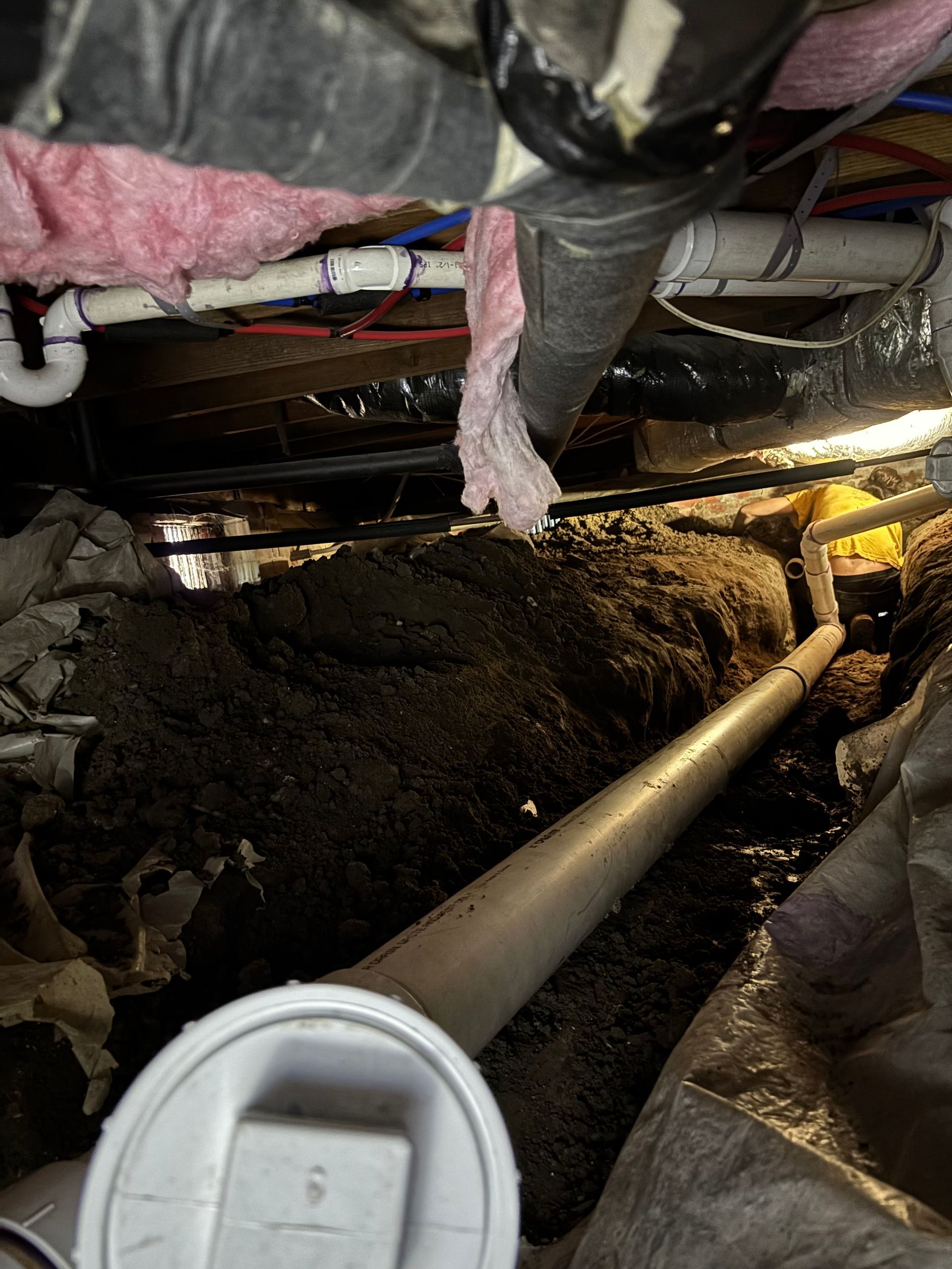 Looking up into a crawl space filled with dirt, pipes, insulation, and structural beams. A long, white PVC pipe is prominent.