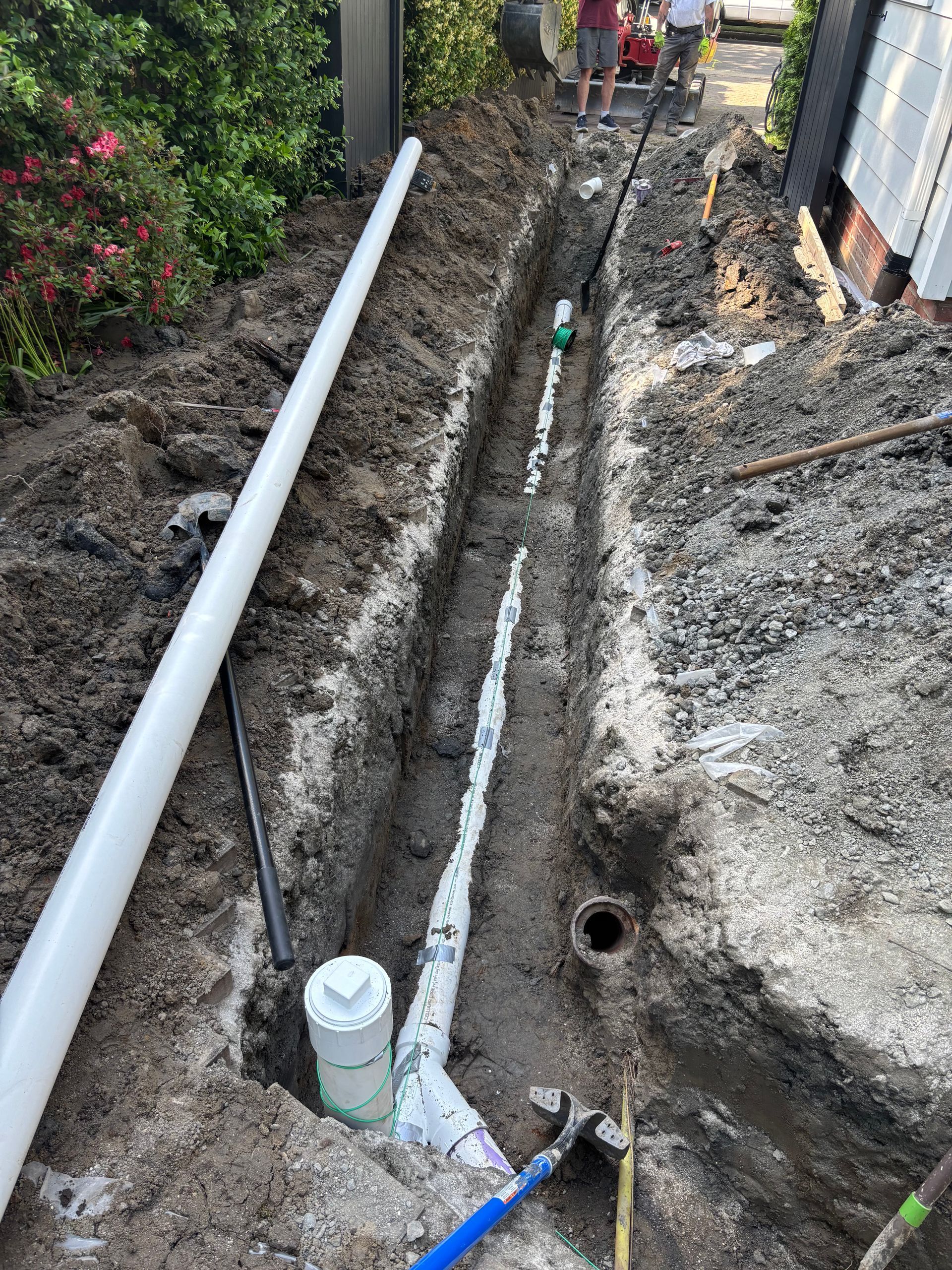 A trench dug outdoors, containing white and black pipes being installed. People are visible in the background.