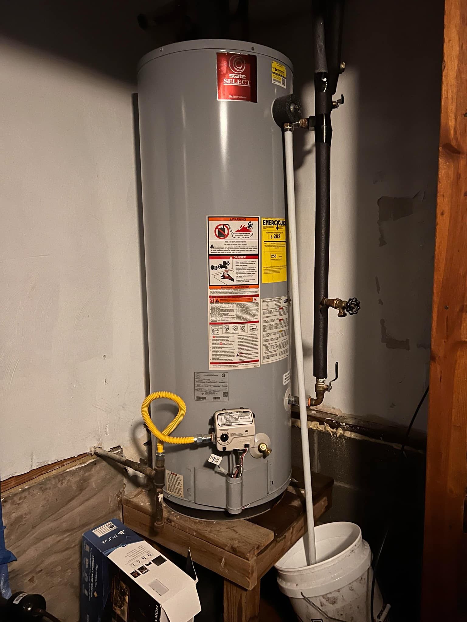A tall, gray water heater stands against a white wall in a dimly lit basement. A yellow gas line connects to the bottom.