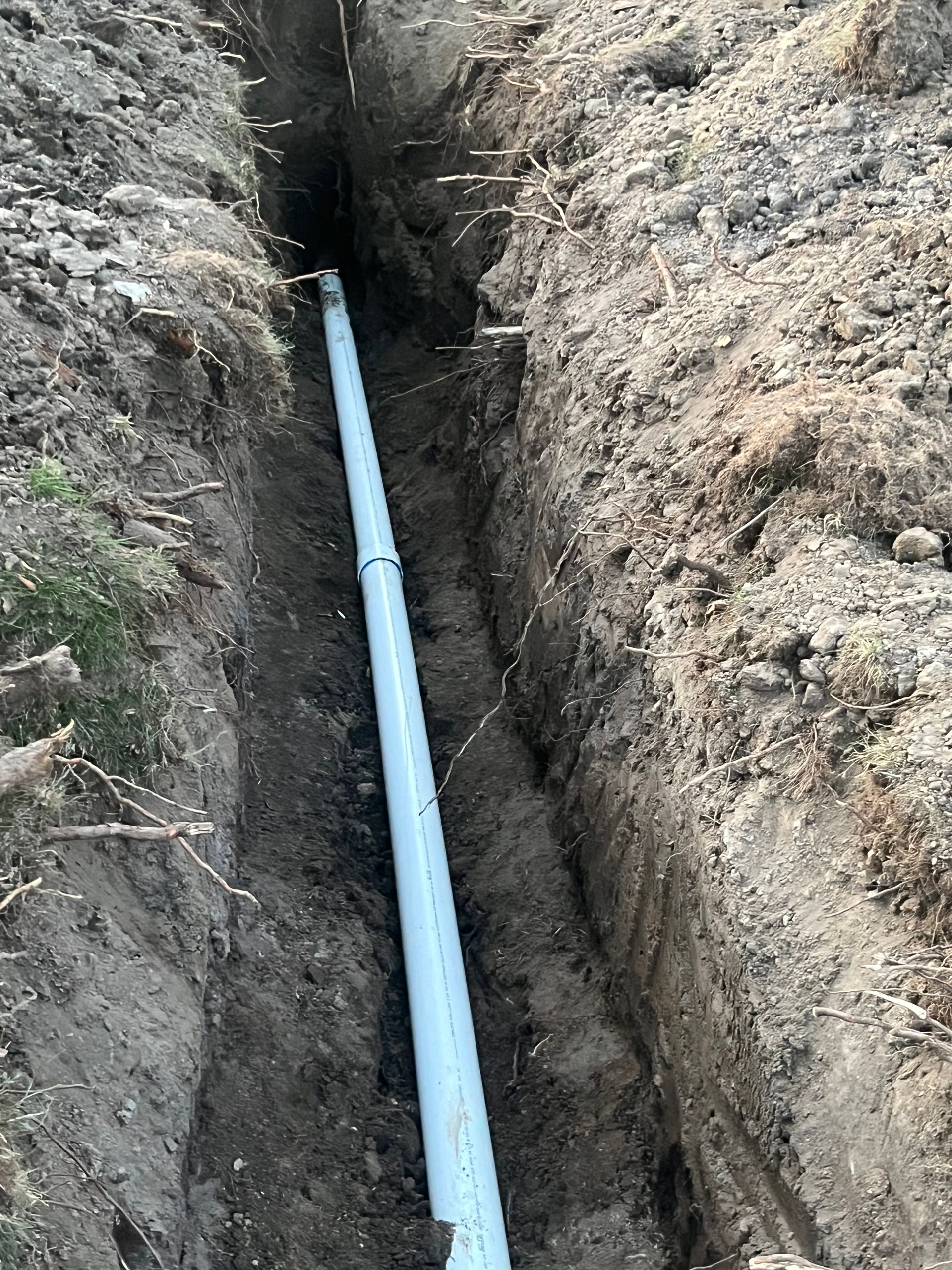 A long, narrow trench with a light blue pipe running through it, likely for utility installation. The setting is outdoors, in dirt.