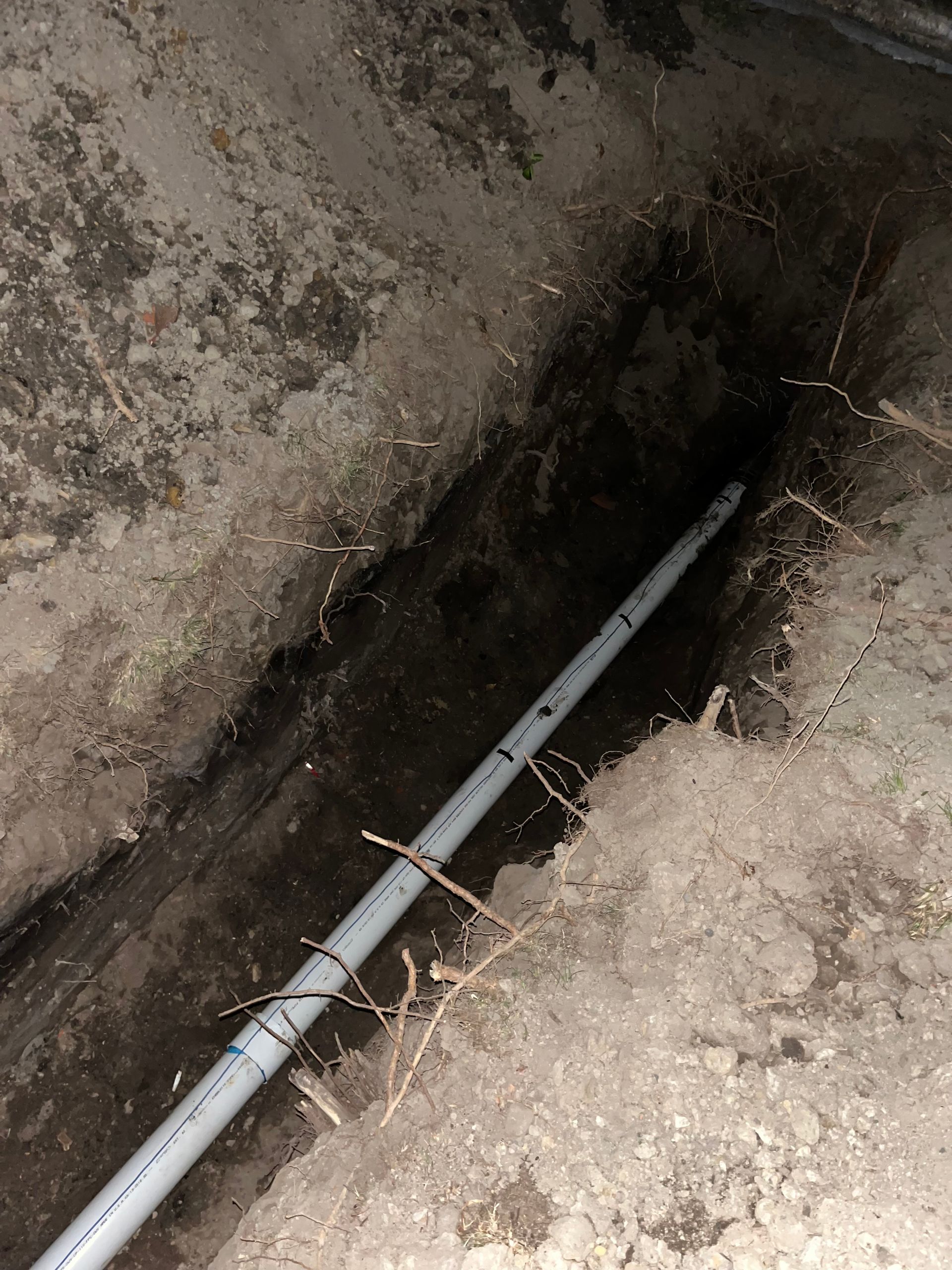 A gray pipe is in a trench dug in the dirt. The trench leads into darkness.