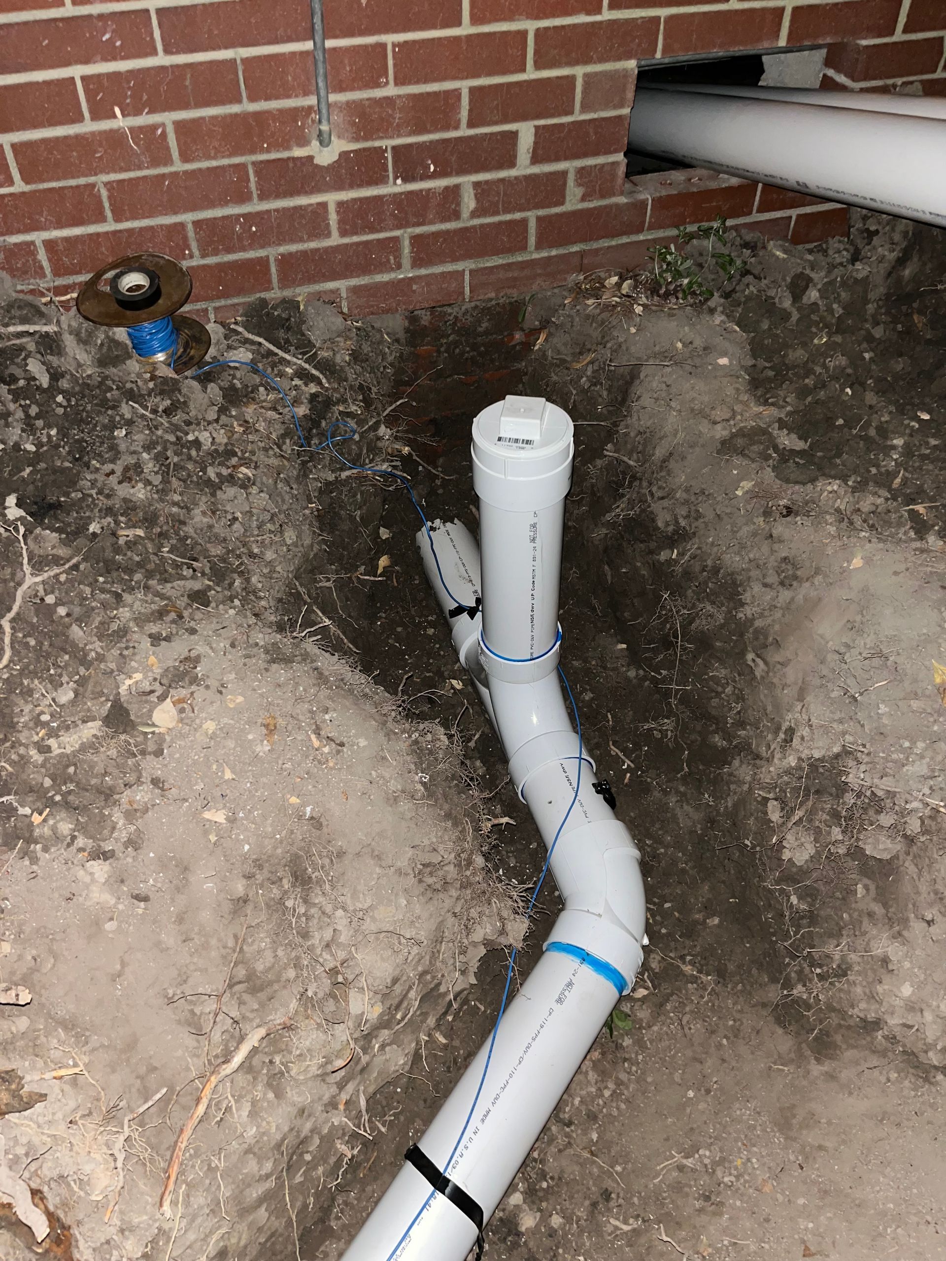 White PVC pipes and fittings partially buried in dirt next to a red brick wall. Pipes extend from a hole and a drain.