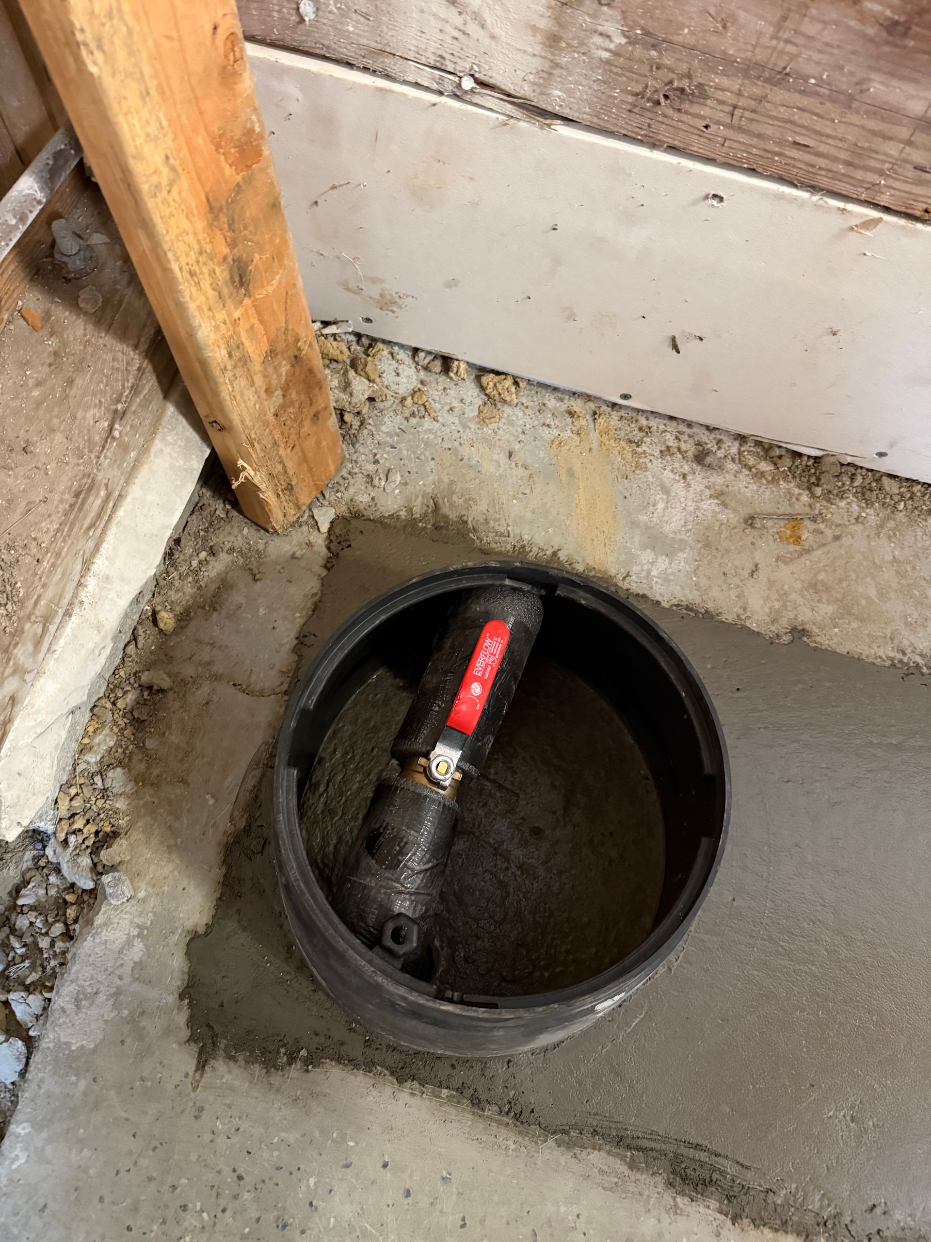Black circular access point with a red light and plumbing in a concrete floor, near wood and a wall.