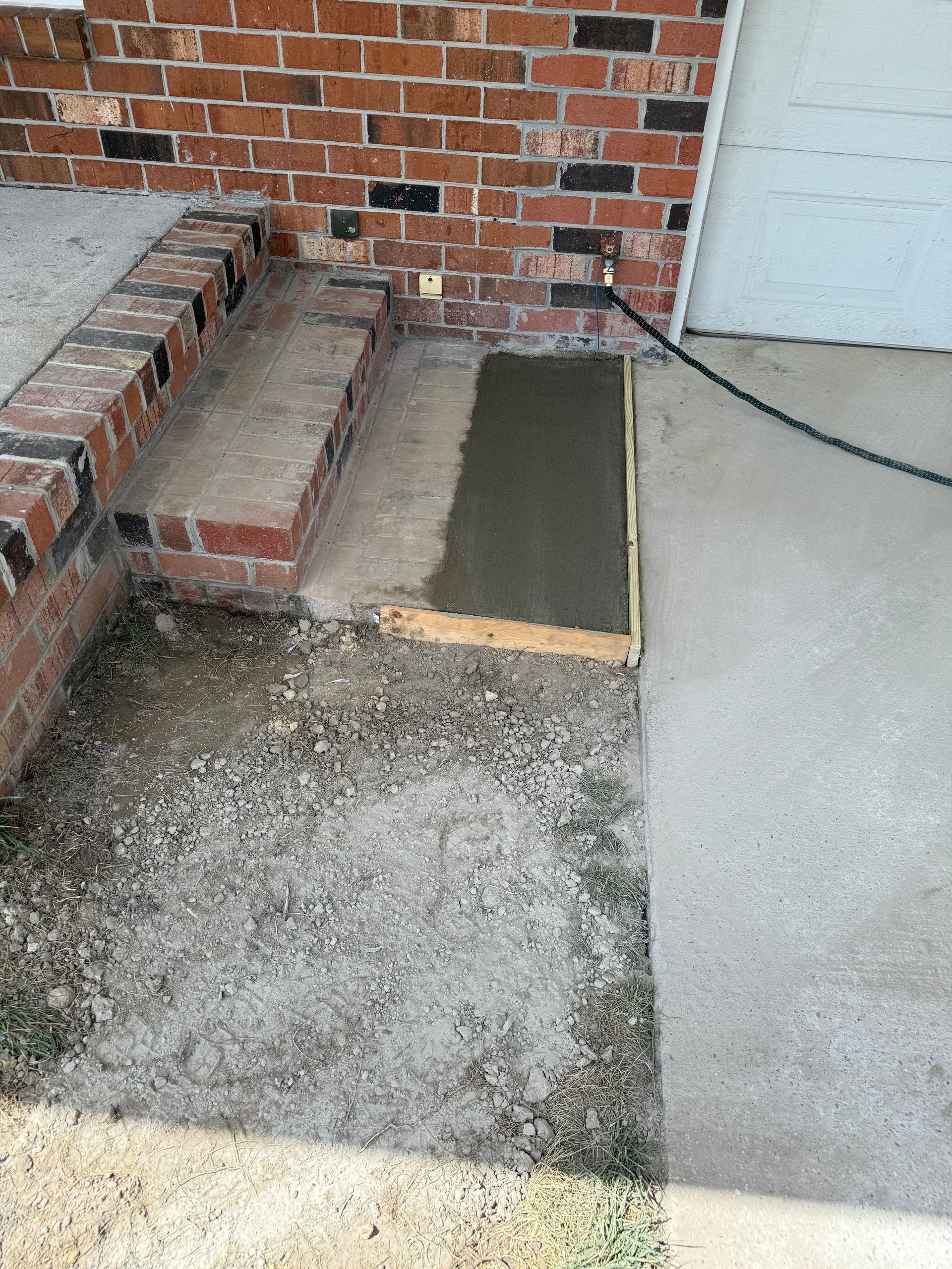 A newly poured concrete slab bordered by wood framing next to steps. The area has gravel and is next to a garage.
