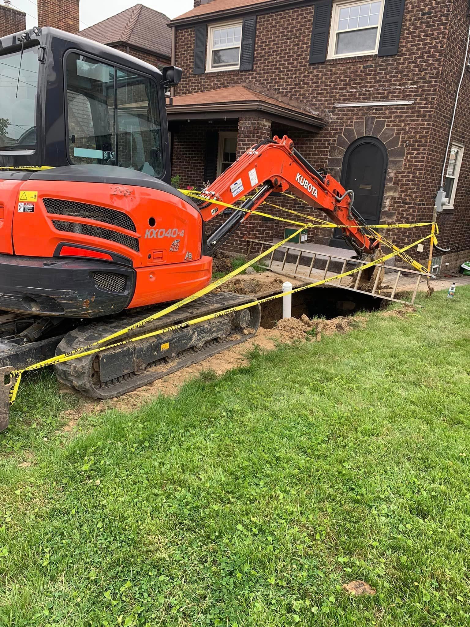 An excavator is digging a hole in the ground in front of a brick house.