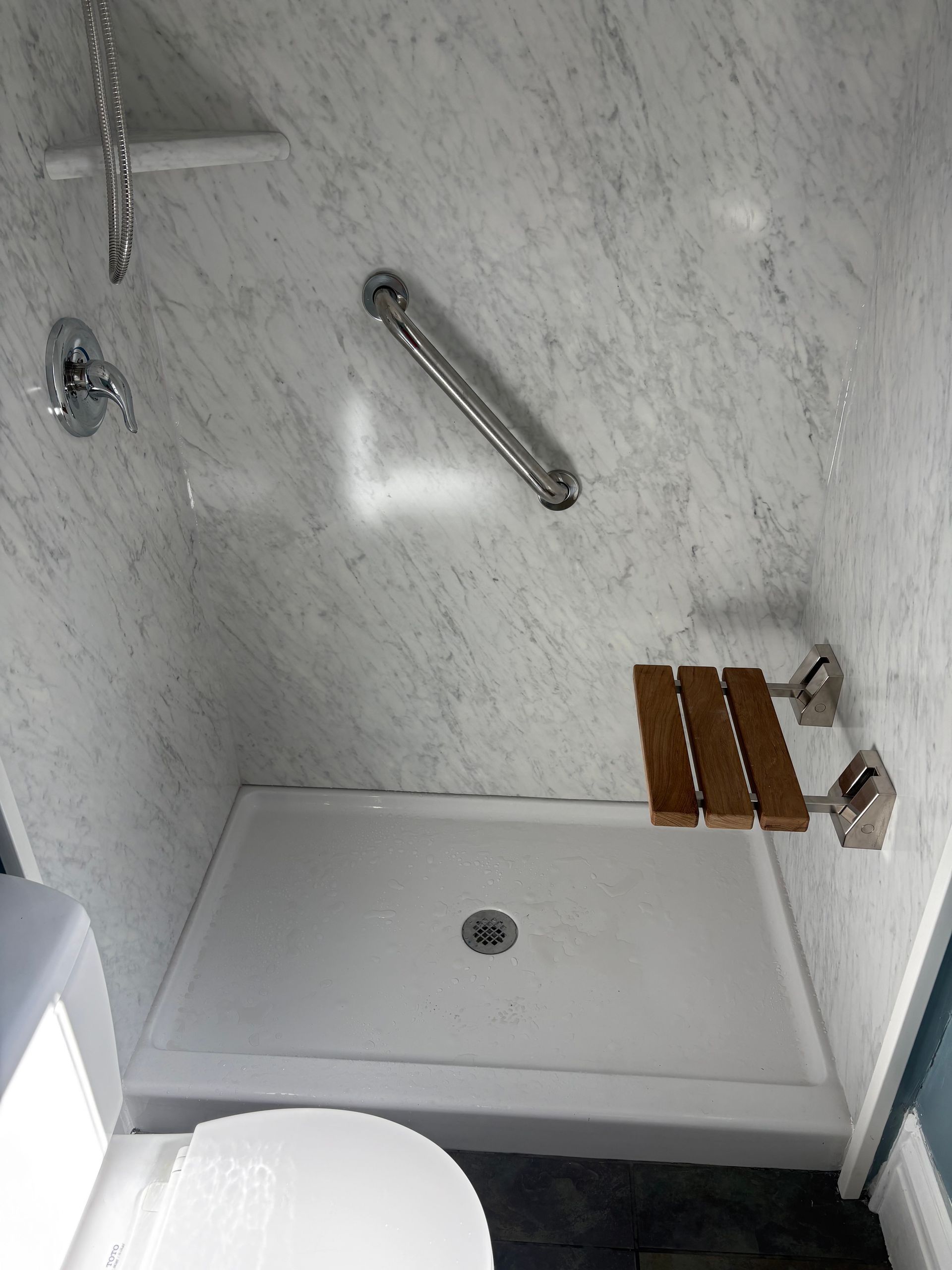 Accessible shower with grab bar and folding wooden seat; white walls, shower base, chrome fixtures.