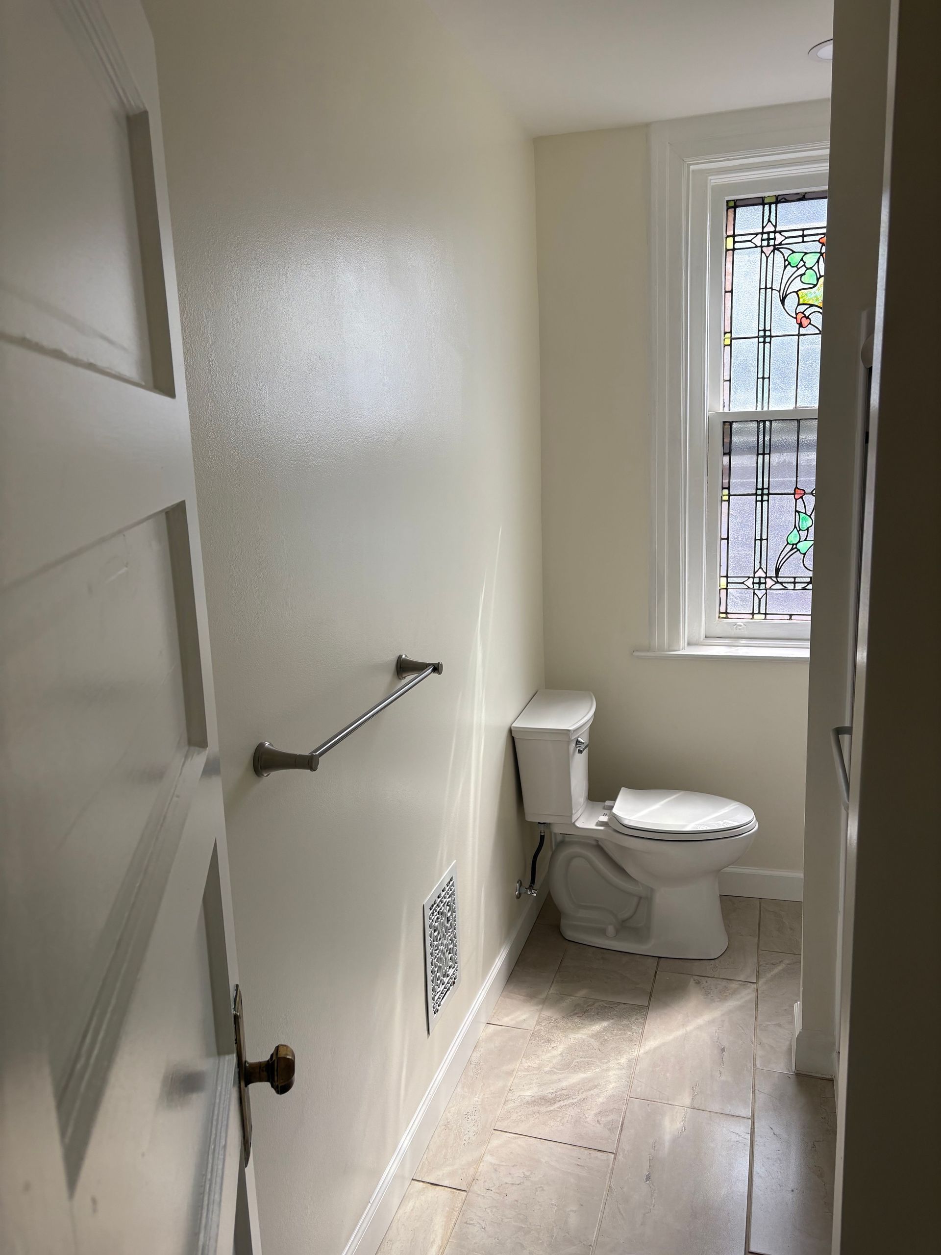 Small, light-colored bathroom with toilet, towel rack, and stained-glass window.