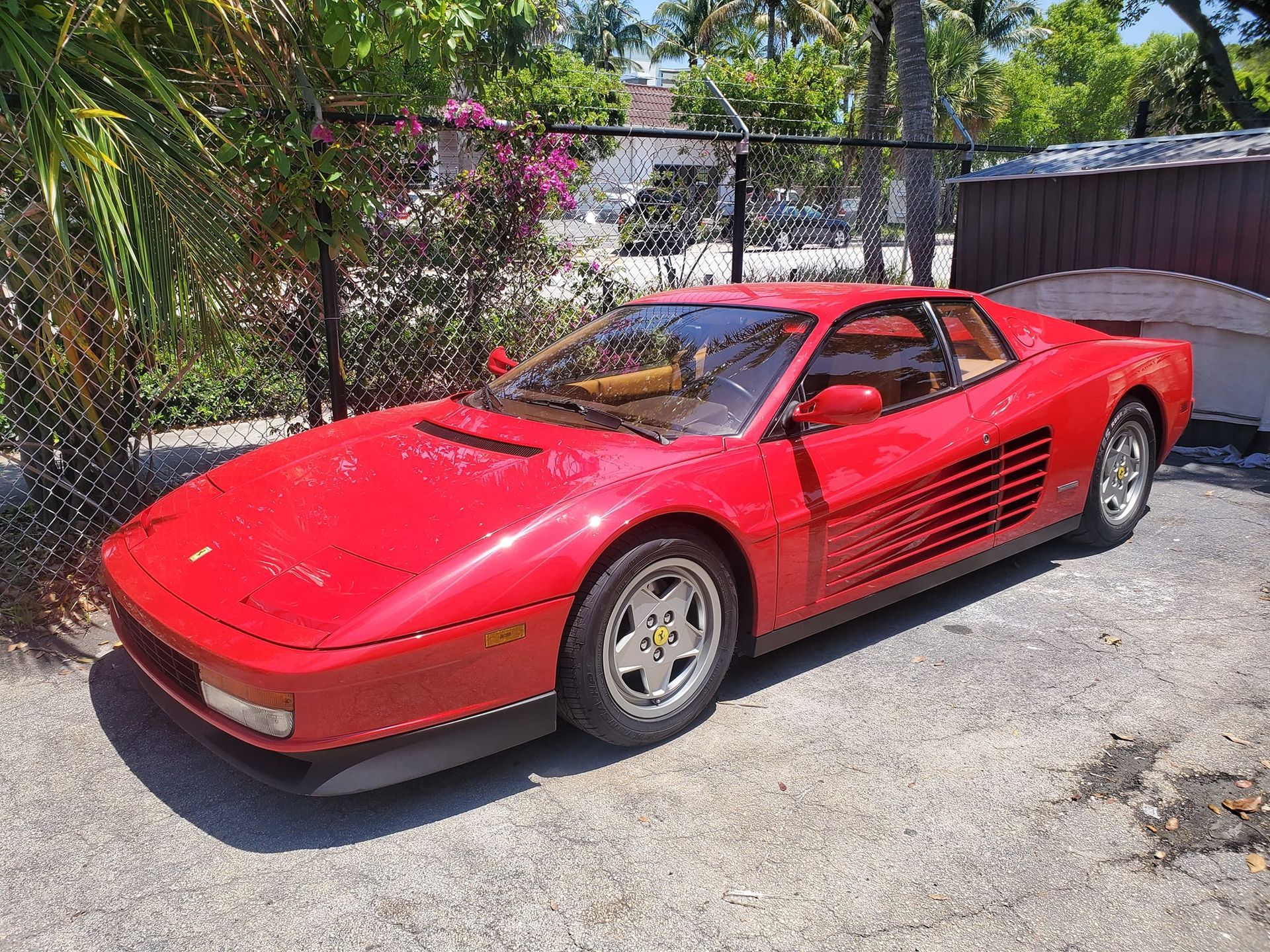 A red ferrari testarossa is parked in a driveway next to a fence.