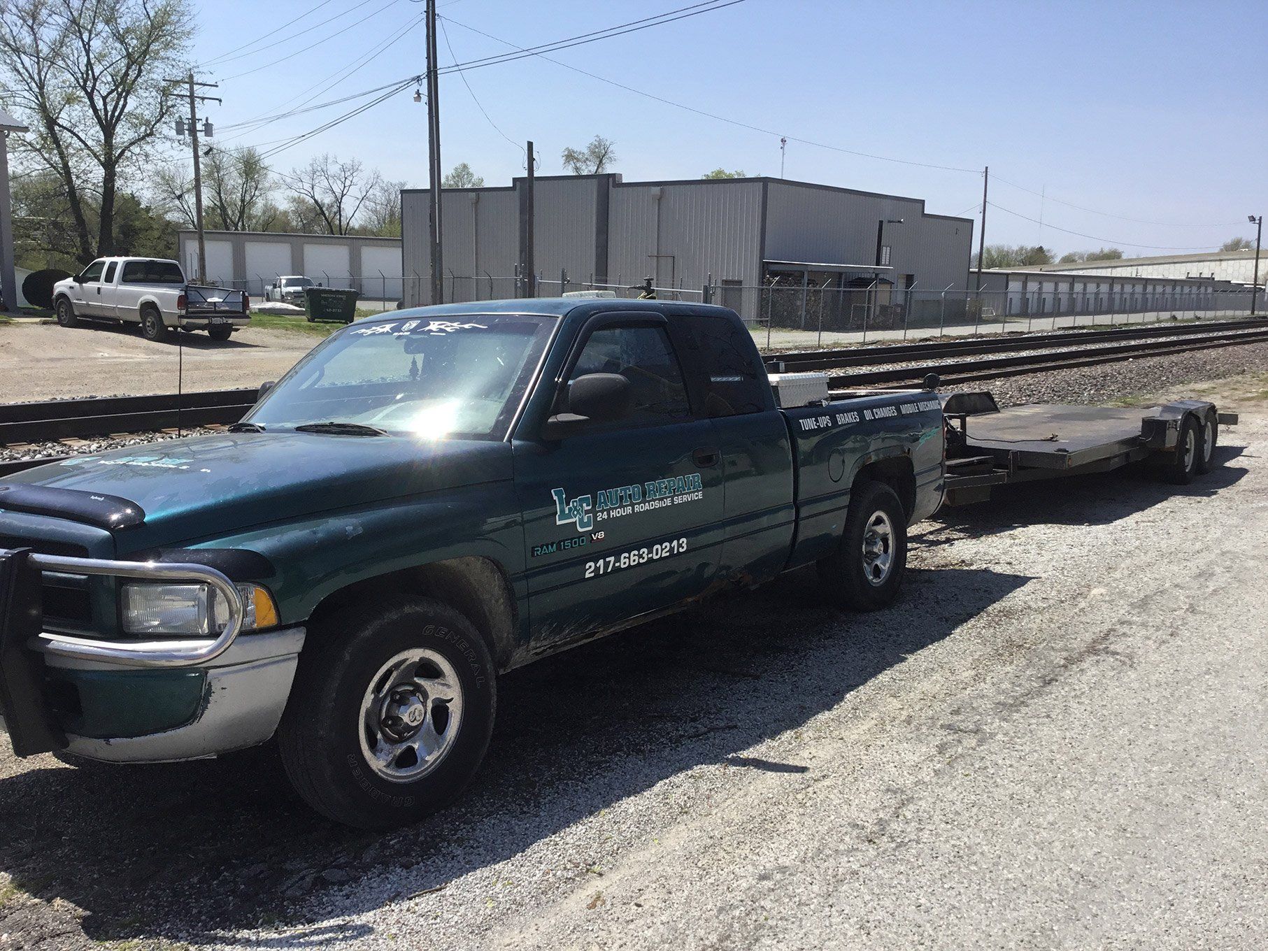 Dark green tow truck with trailer parked on gravel lot near industrial buildings and train tracks